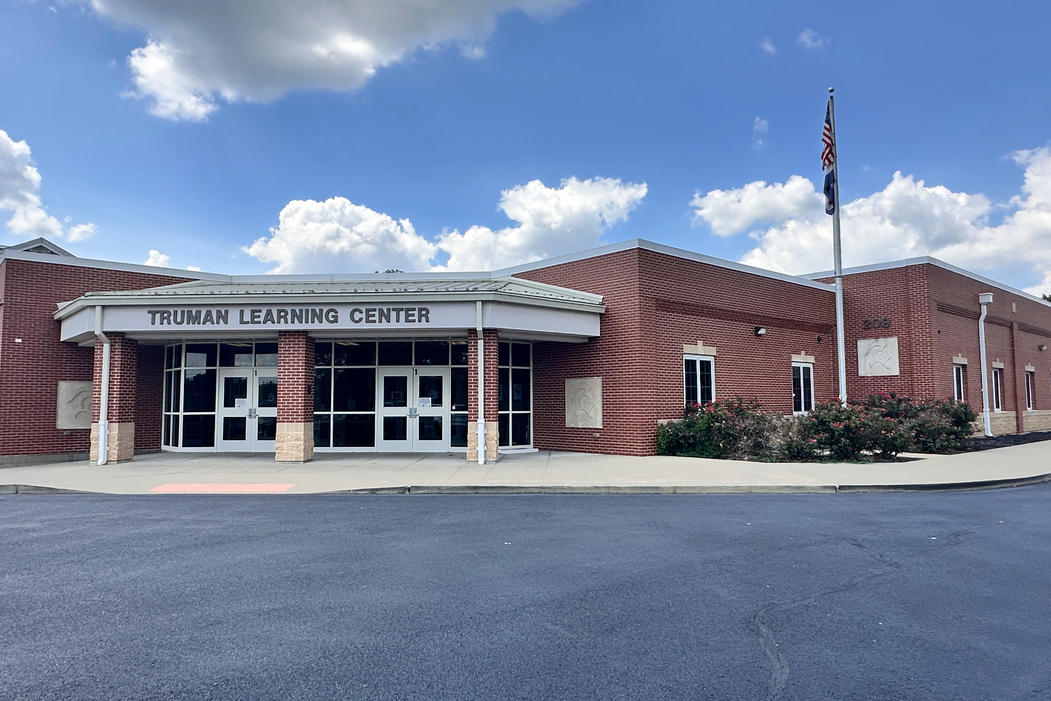 Exterior of a brick building with "Learning Center" sign. A flagpole stands by the entrance, which has glass doors. Trees and plants line the sidewalk.