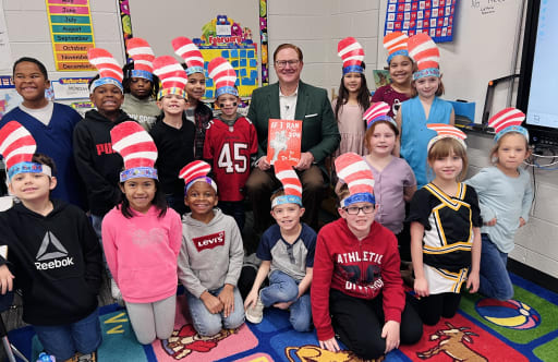 Superintendent With students reading a book at library all with hats of dr seuss