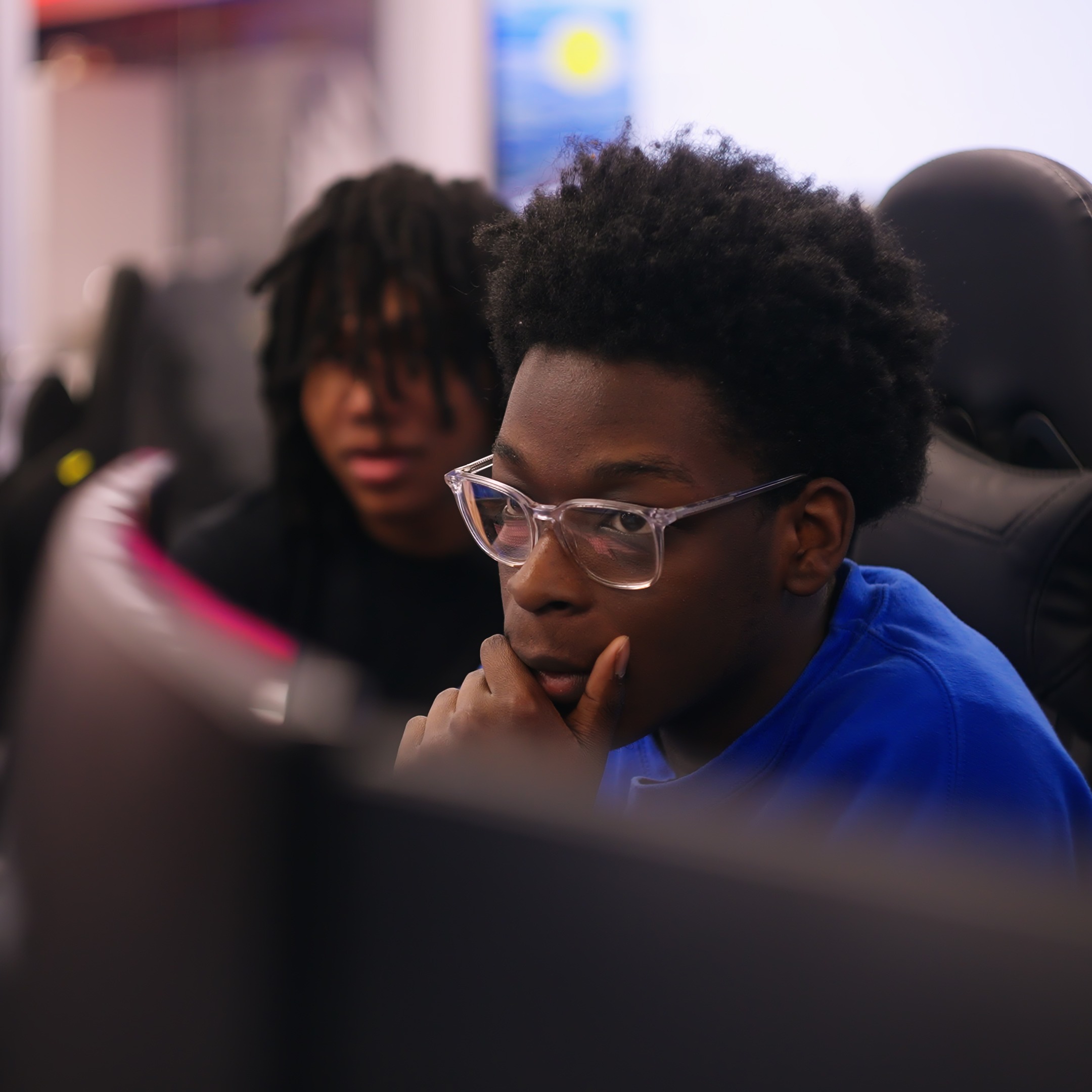 Two individuals seated at computer monitors, one with glasses and a blue shirt, the other in a black shirt.