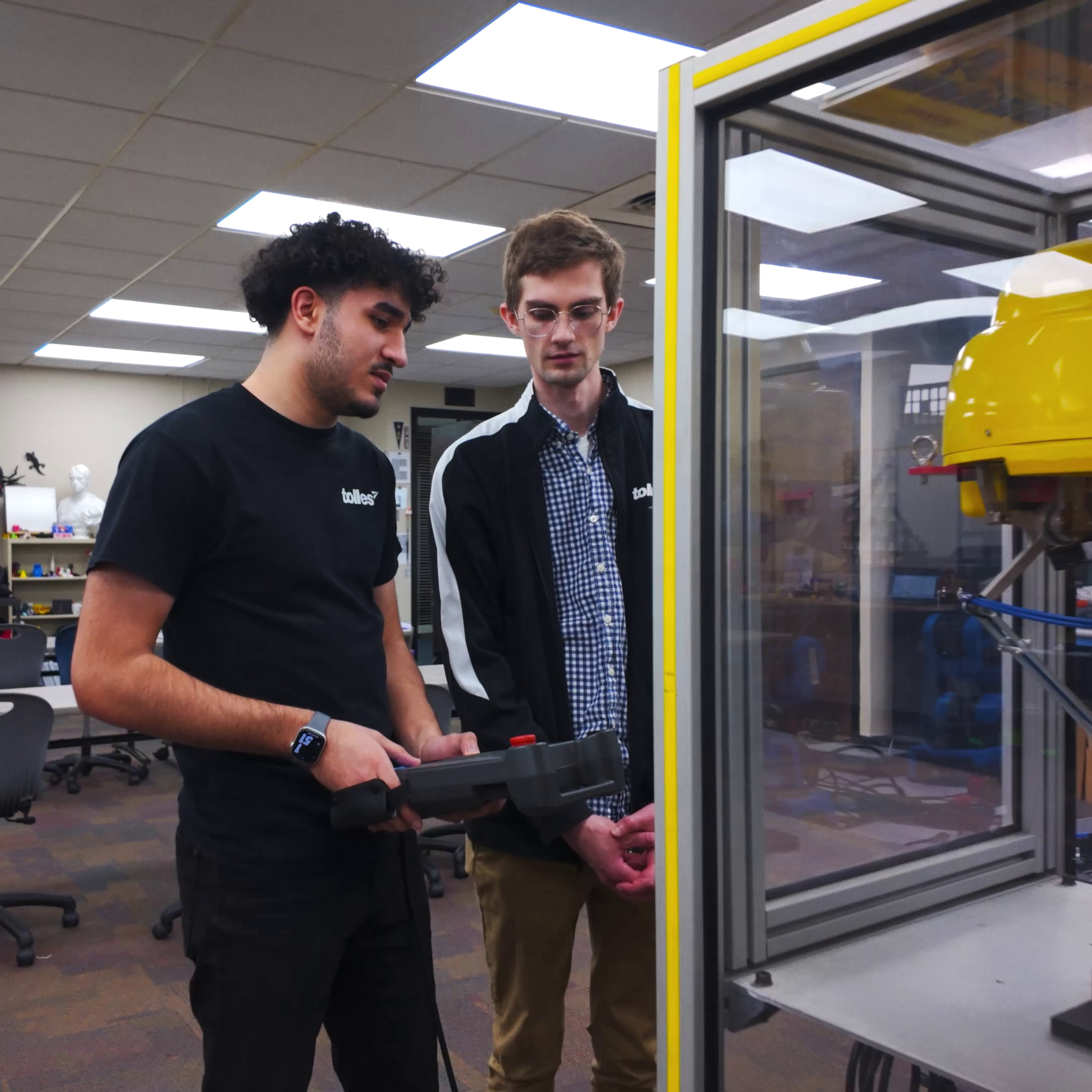 Two people in a lab with a yellow machine, one holding a device and the other wearing a watch.
