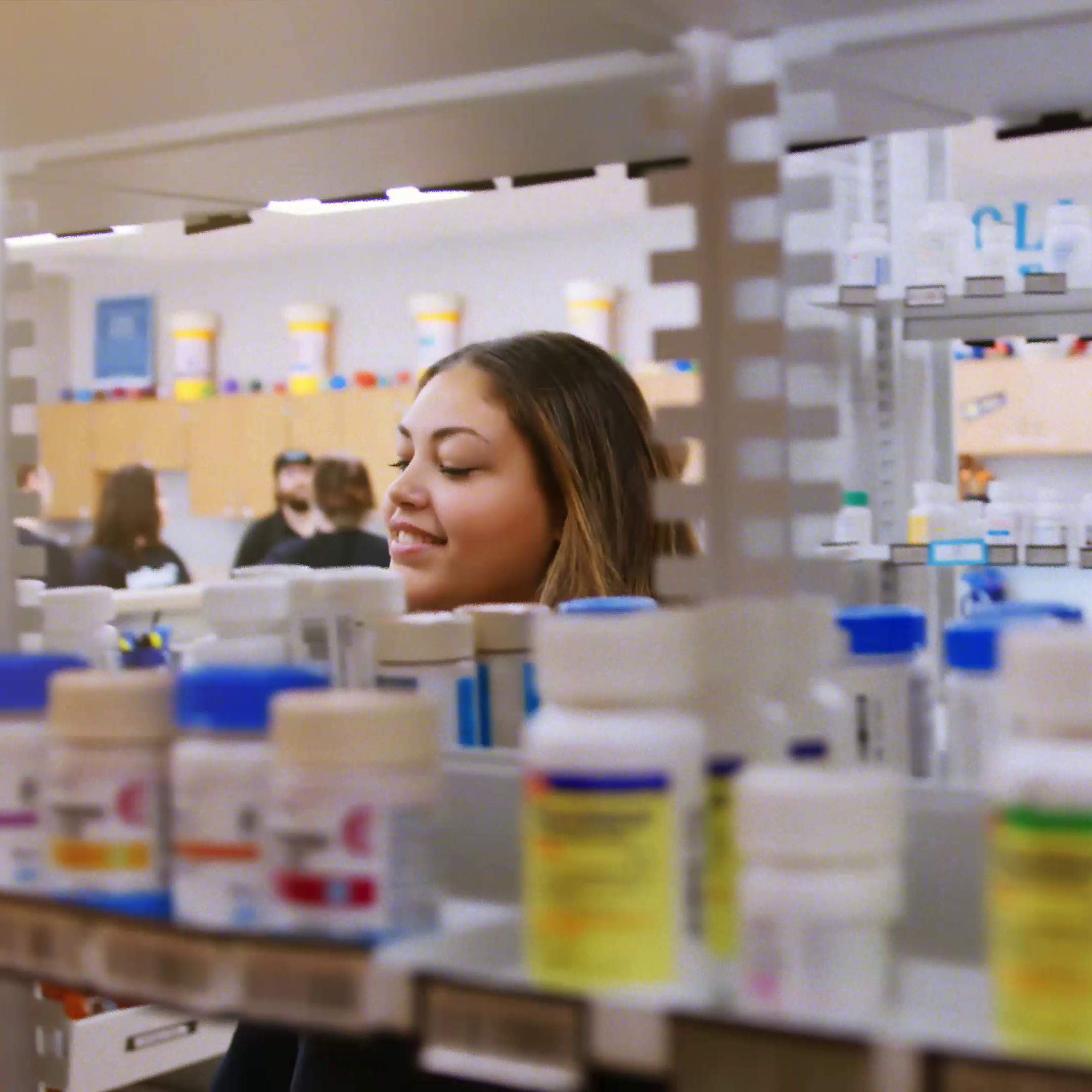 A woman smiles in a pharmacy, looking at shelves filled with various medications. Background shows blurred figures and pharmacy shelves.
