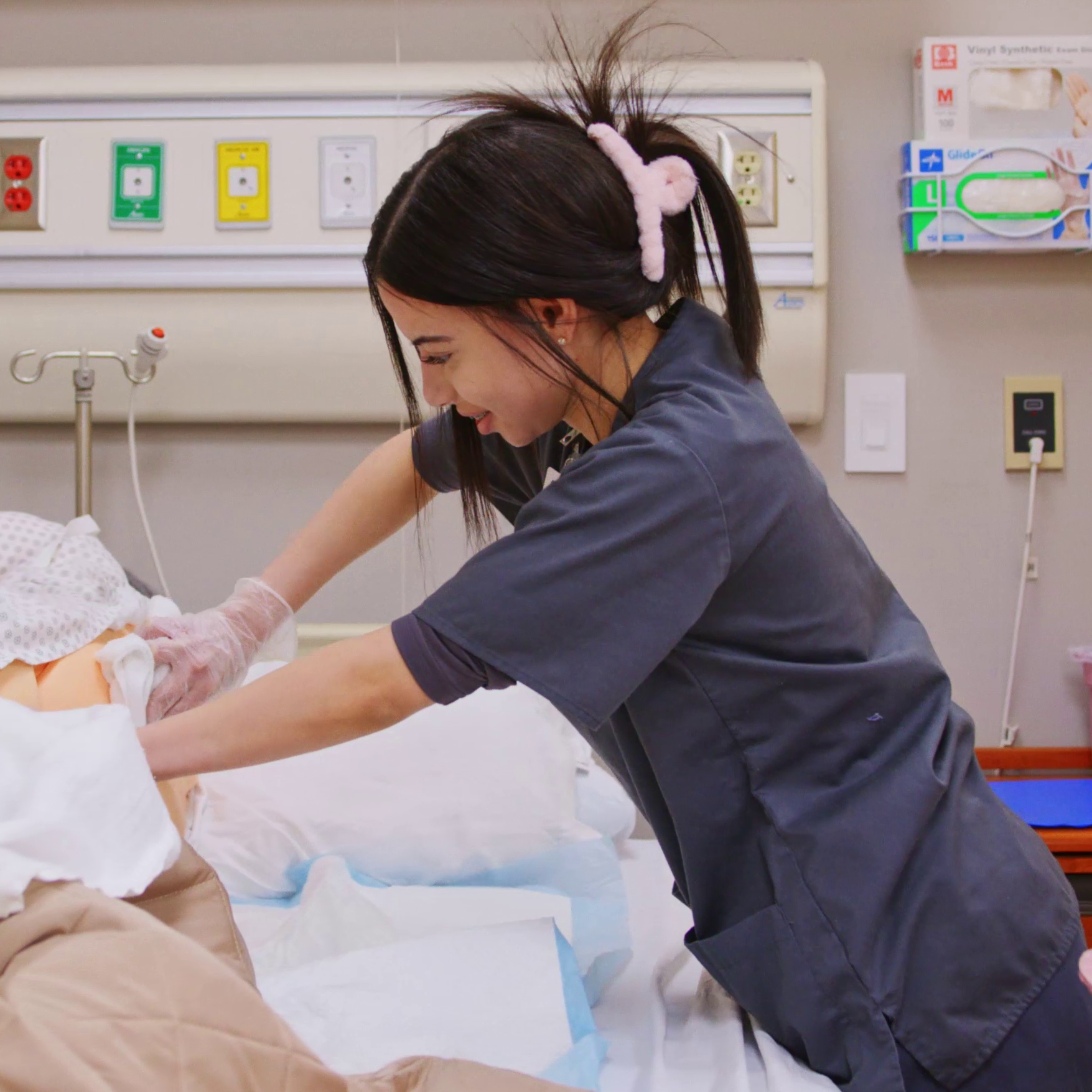 A person in scrubs is in a hospital room adjusting a mannequin's sheet on a bed.