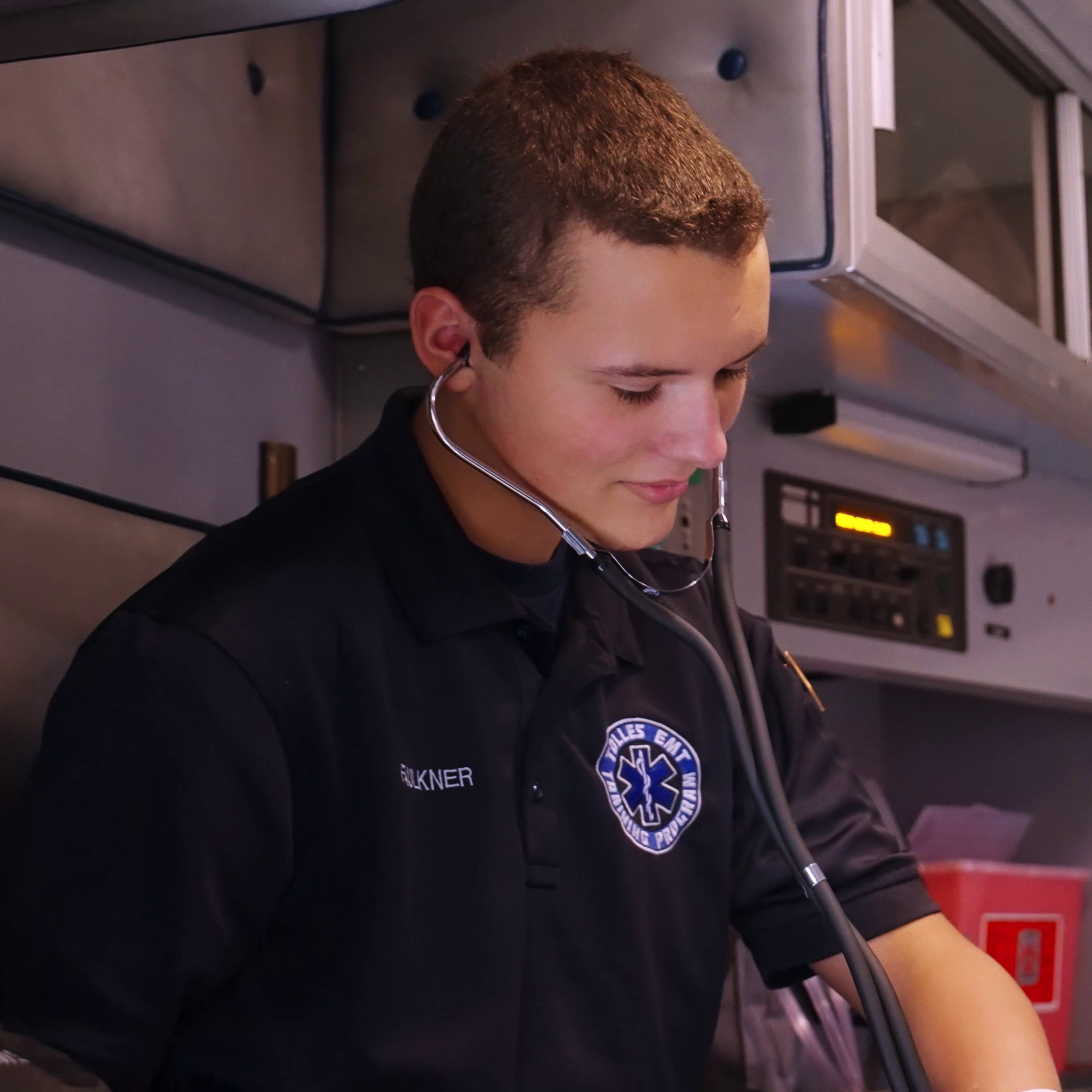 A man in uniform with a stethoscope around his neck is seated inside an ambulance, listening to a patient.