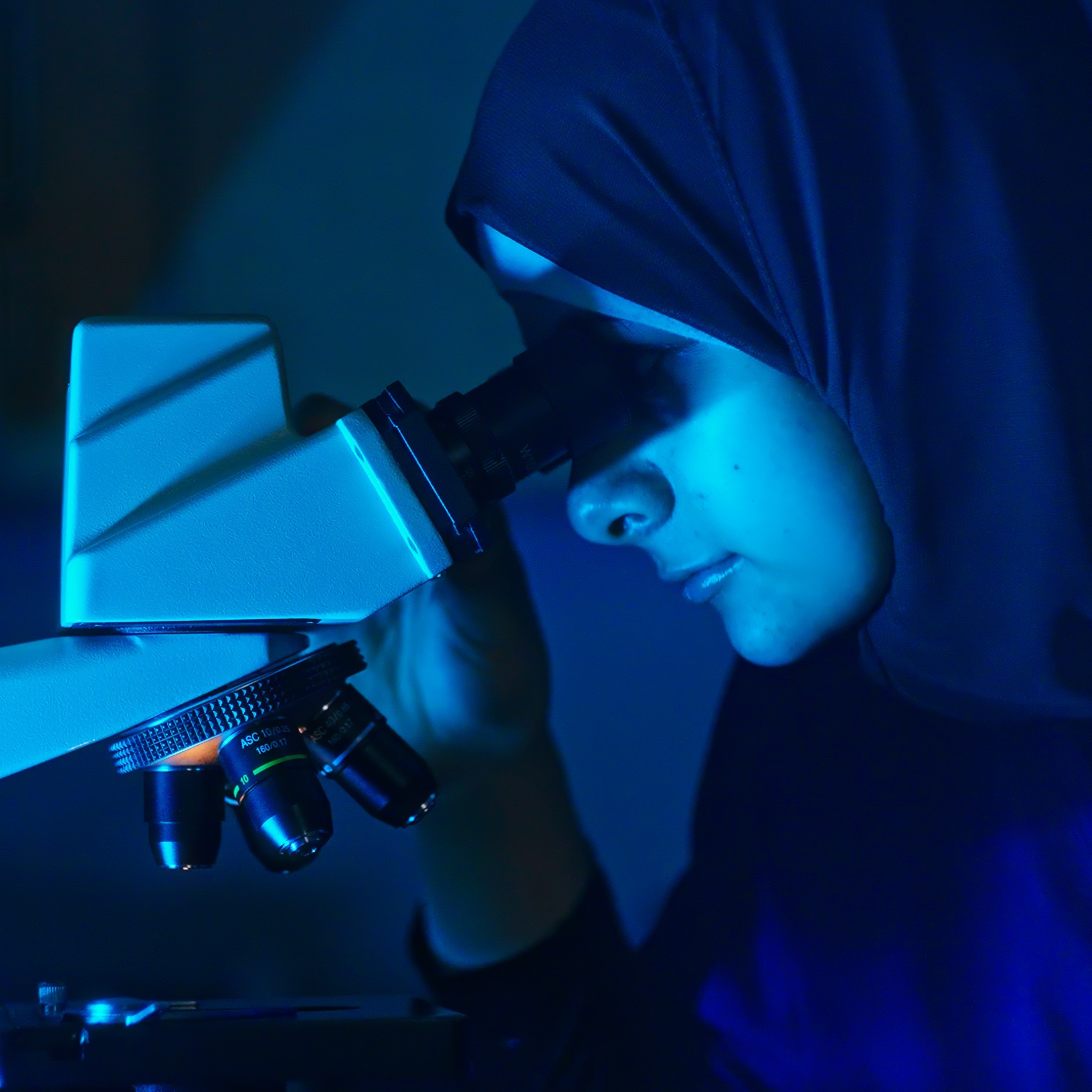 Person in a blue hijab looking into a microscope with blue light illuminating the scene.