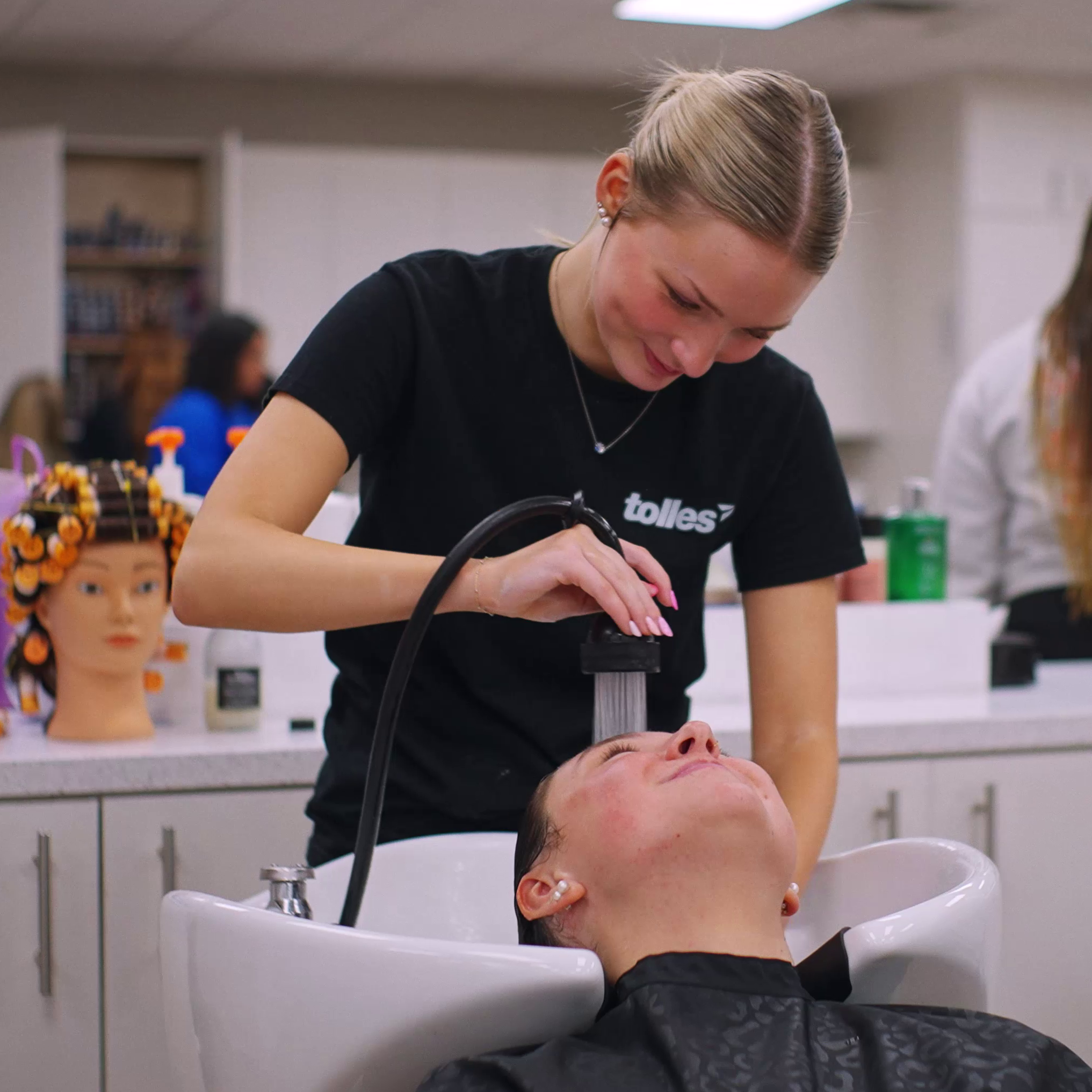 A woman shampoos a man's hair in a salon with other people and a white desk in the background.