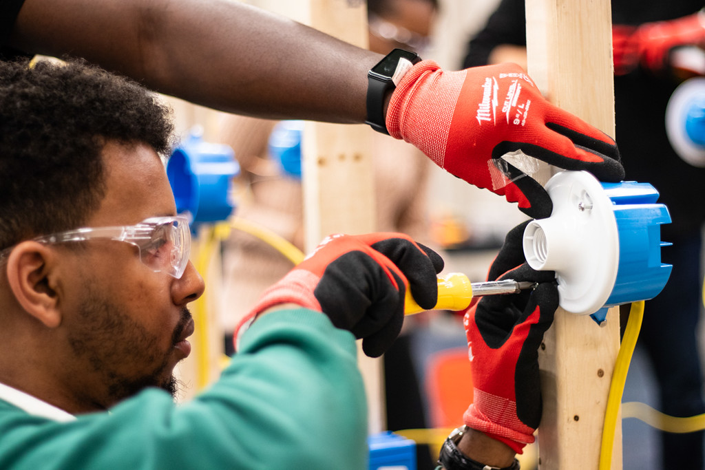 A man in green and red gloves works on a blue and white device, holding a screwdriver, while another person assists.