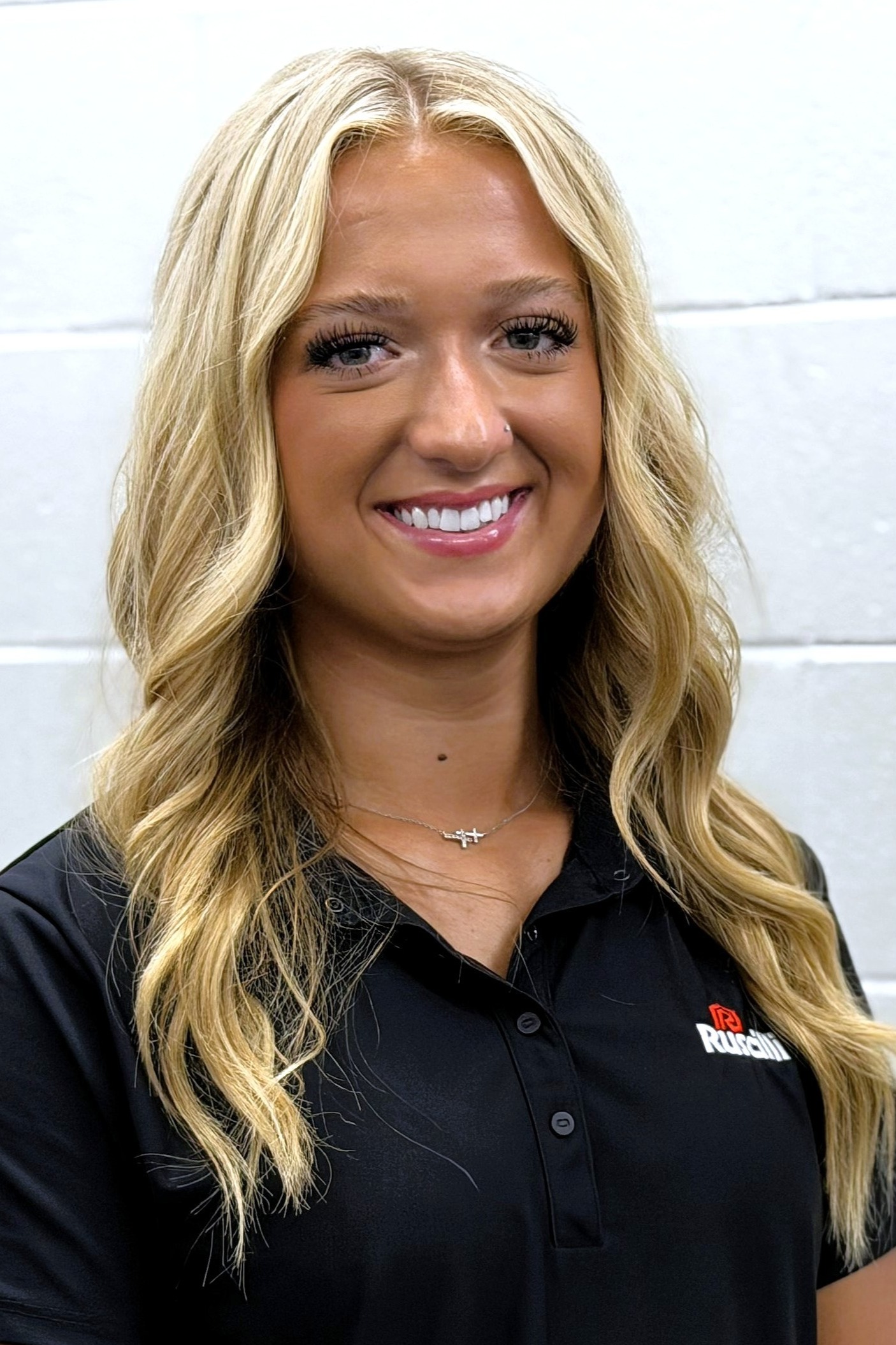 A woman with blonde hair and a black polo stands against a white wall, smiling at the camera.