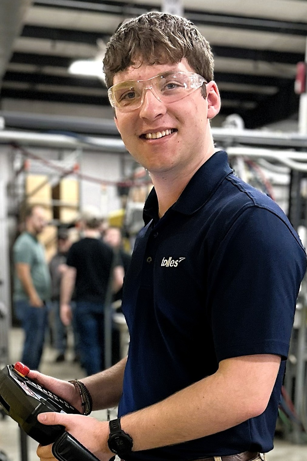A man in a blue shirt and khakis stands in a factory, holding a tool and standing next to a yellow machine.