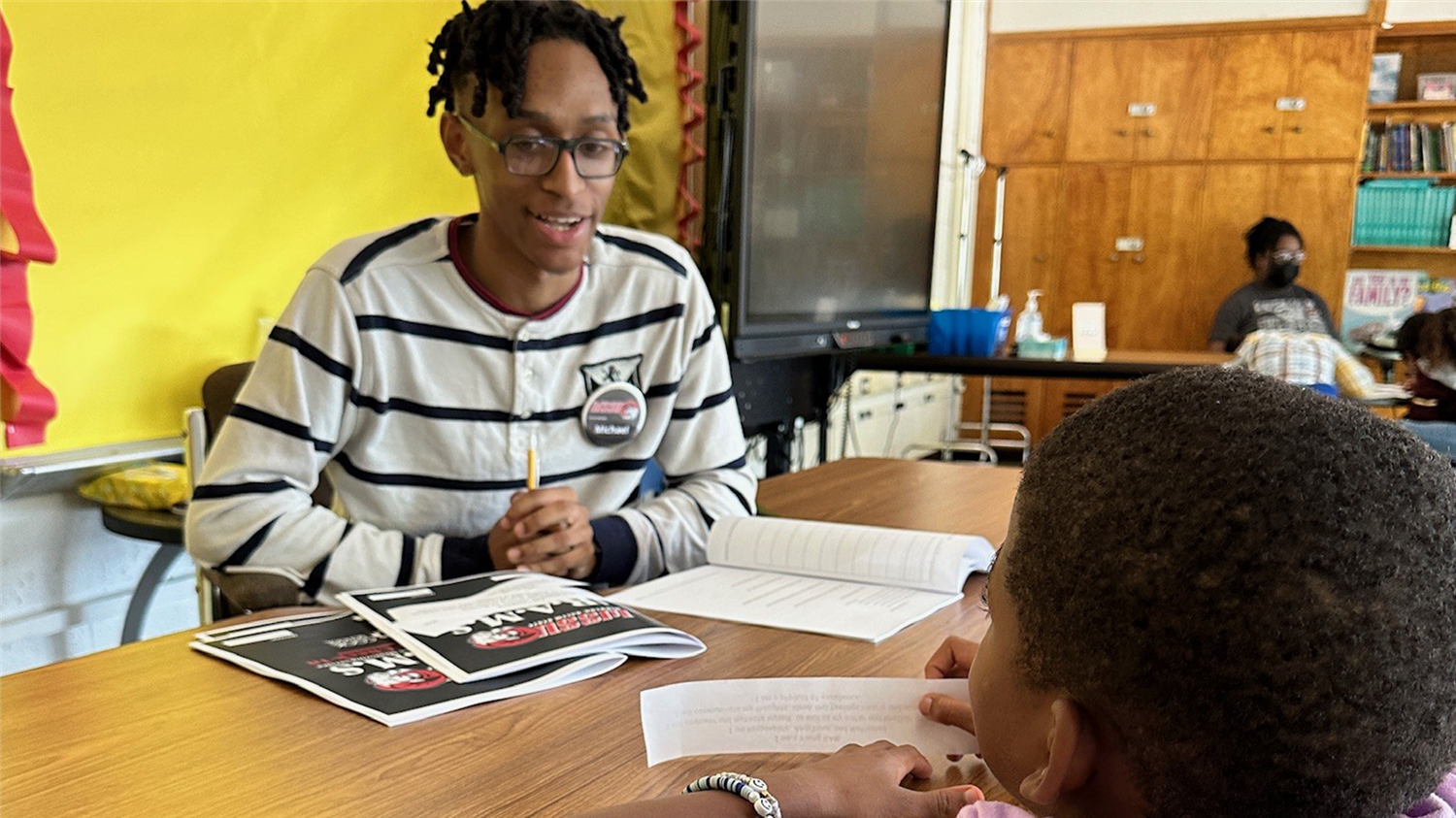 A young man seated at a table with books and papers, engaged in a conversation or activity with a child.