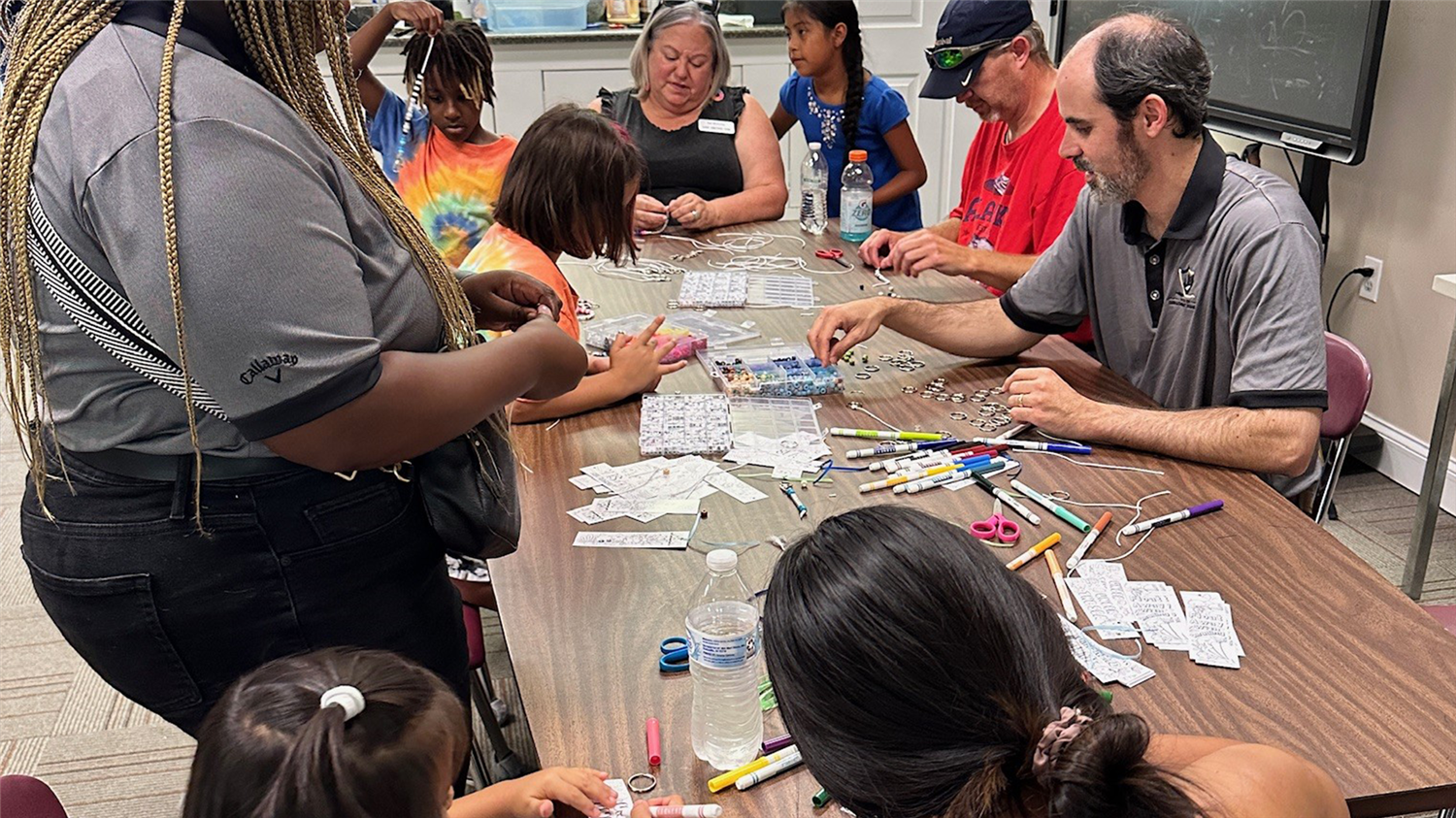 People gathered around a table filled with art supplies, engaged in a creative activity. A person is demonstrating a craft to others.