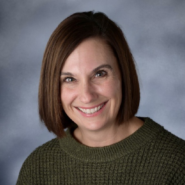 A woman with short brown hair smiles at the camera. She wears a green knit sweater against a solid blue background.