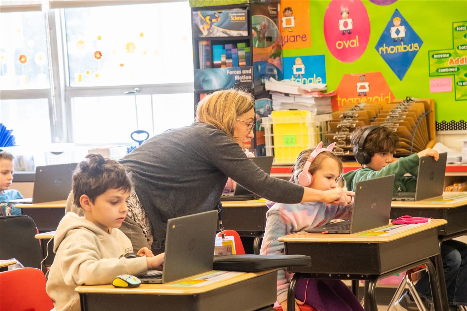 kids in a classroom with computers and a teachir pointing out at one computer of a kid