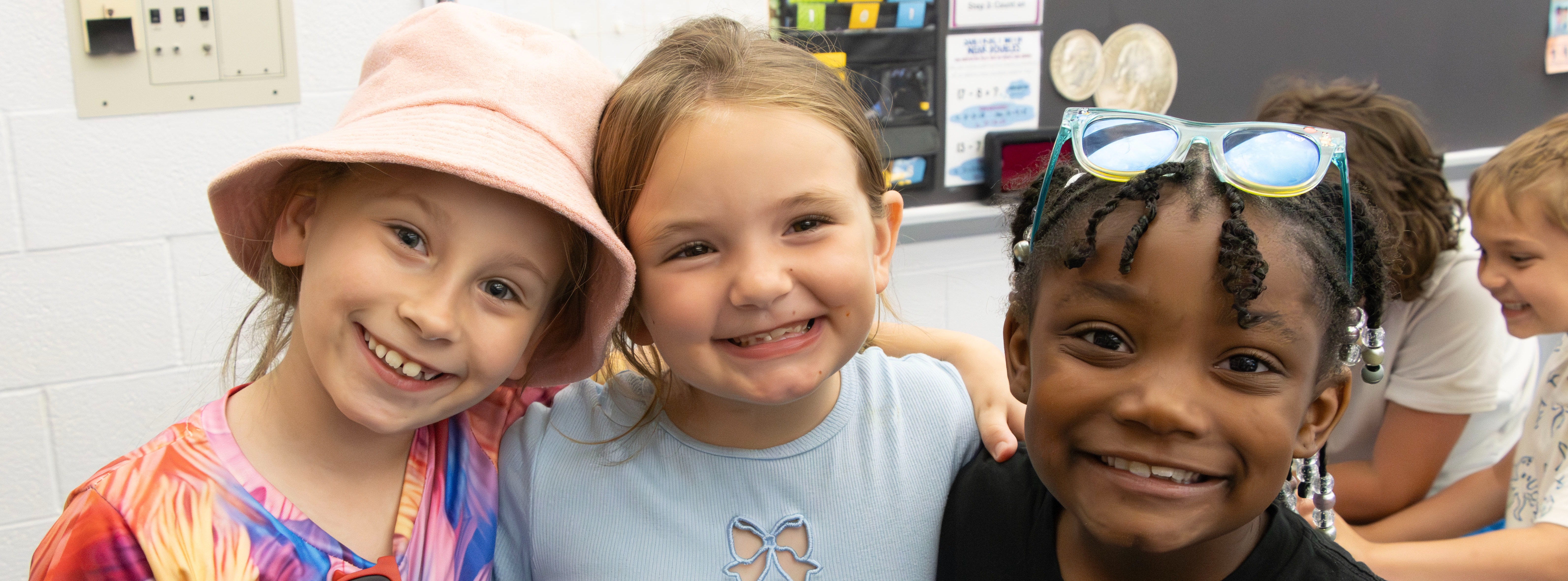 Three young students stand with their arms around one another, smiling at the camera in a classroom. The students are dressed in colorful, casual clothing and accessories while other students participate in activities in the background.