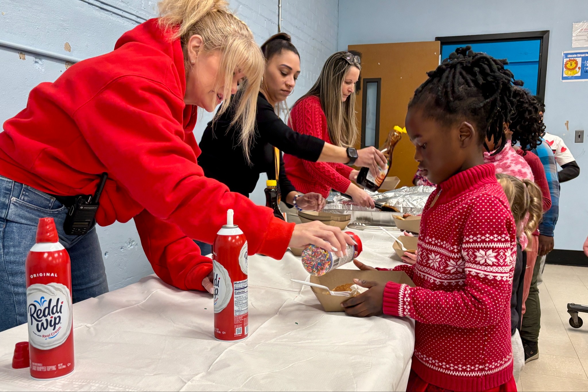 A woman in a red sweater adds sprinkles to a child's dessert at a community event.