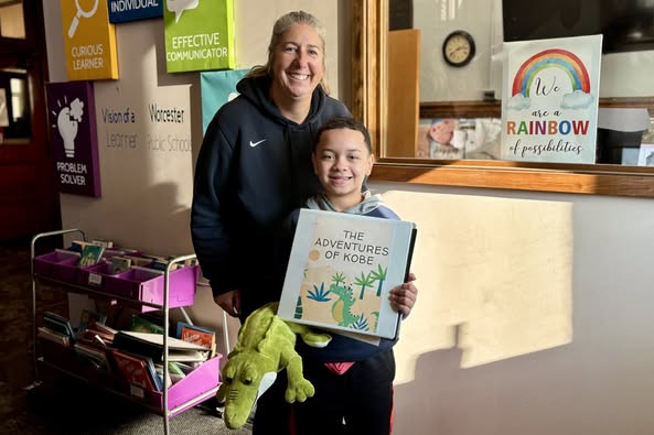 A young student proudly holds a book titled 'The Adventures of Kobe' while standing with an adult in a school hallway.