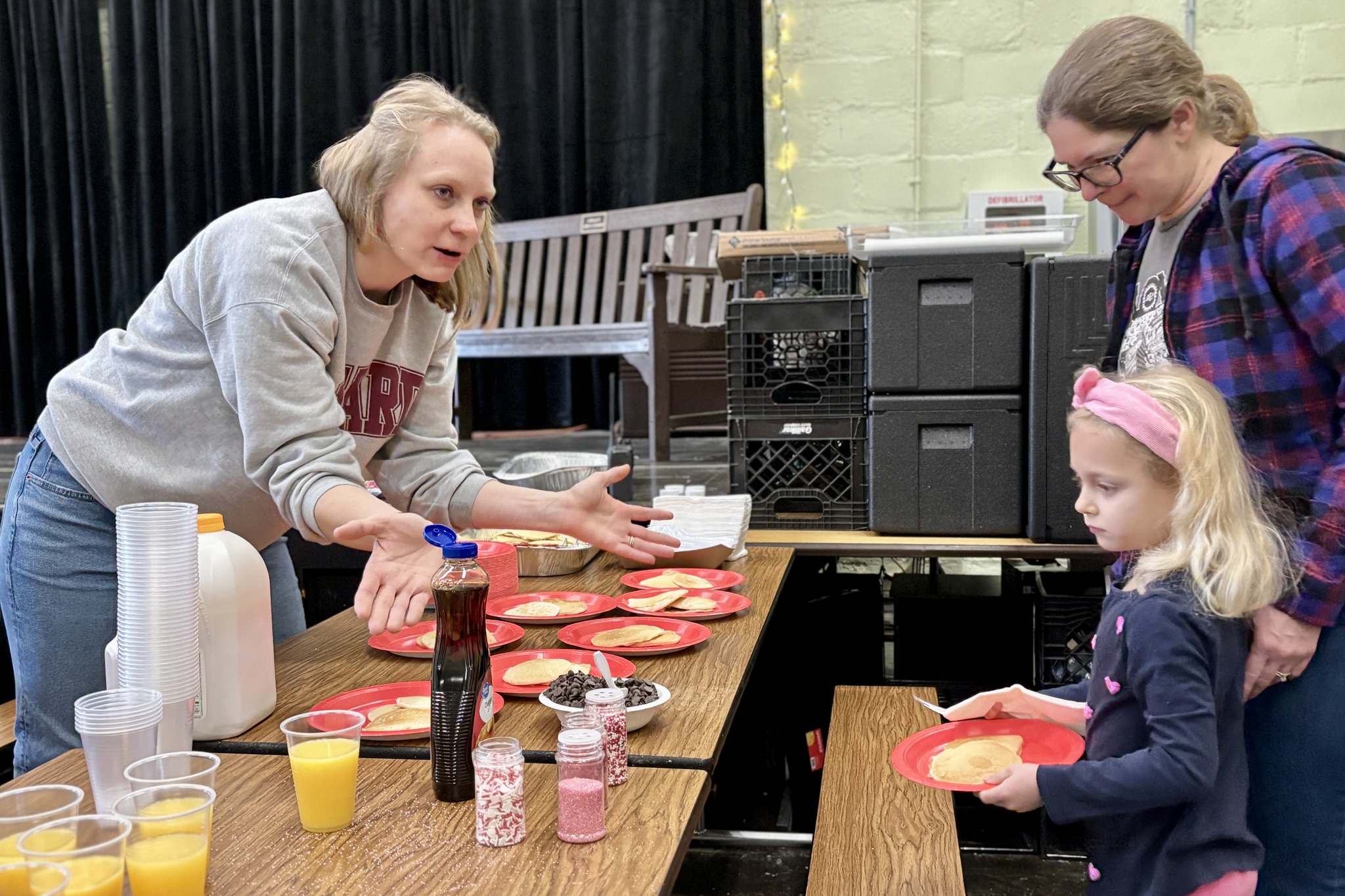 A woman offers a pancake to a young girl at a pancake breakfast.