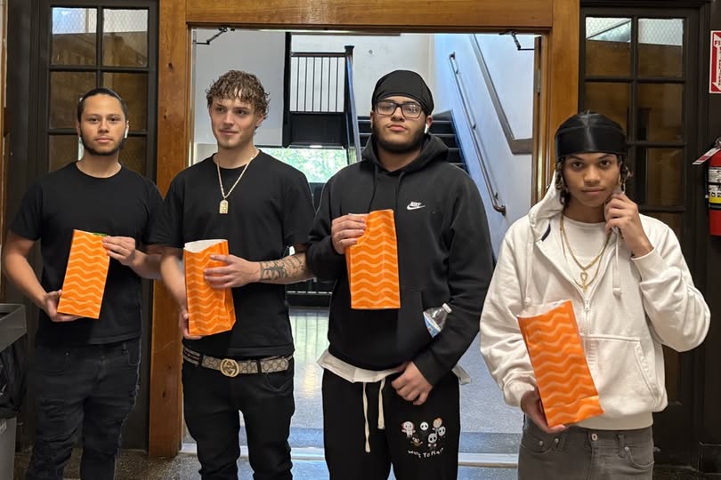 Four young students stand in a hallway, each holding an orange bag with a wavy pattern.