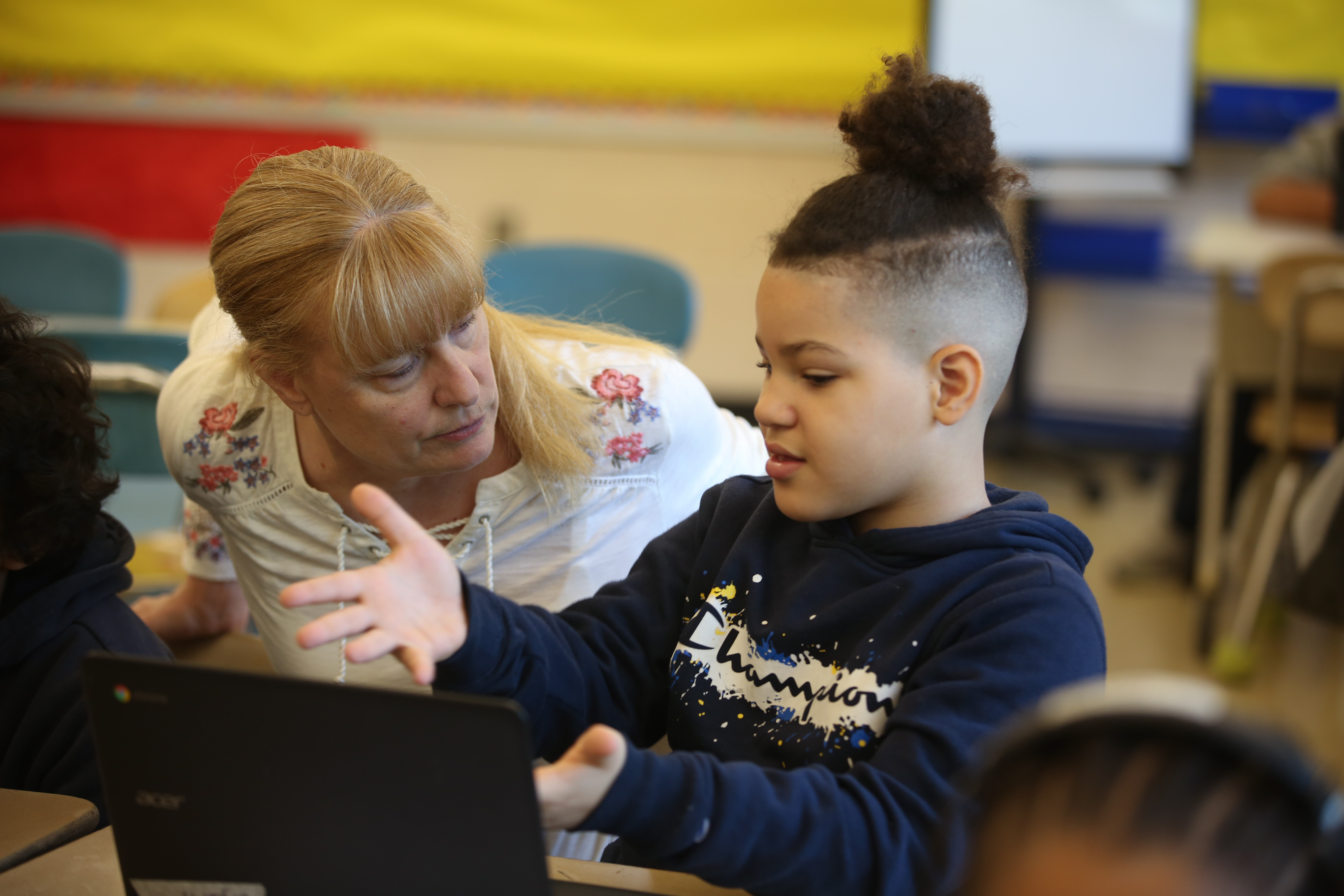 This is a photo of an elementary student talking with their teacher and using technology
