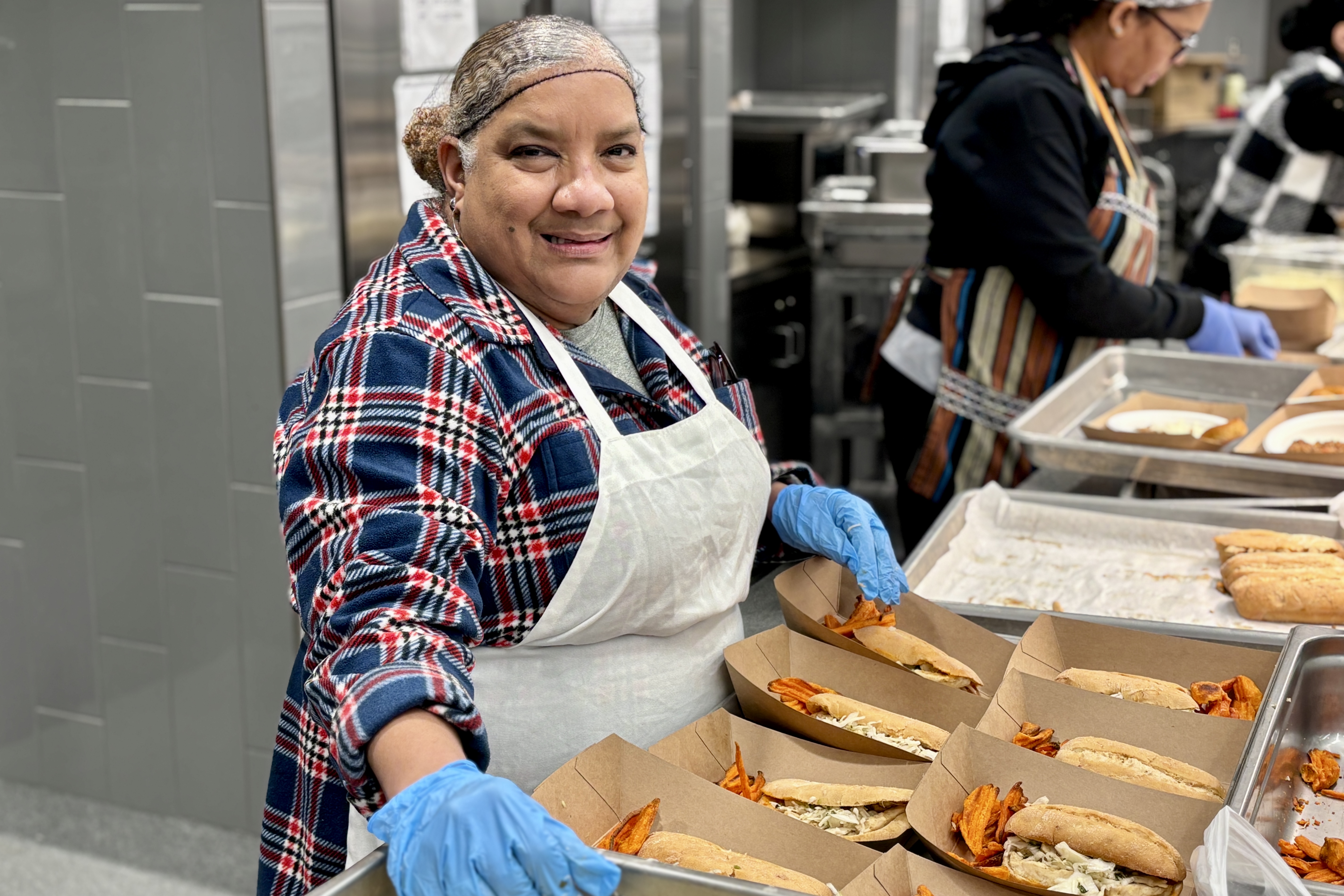 A smiling woman in a kitchen prepares food, wearing gloves and an apron.