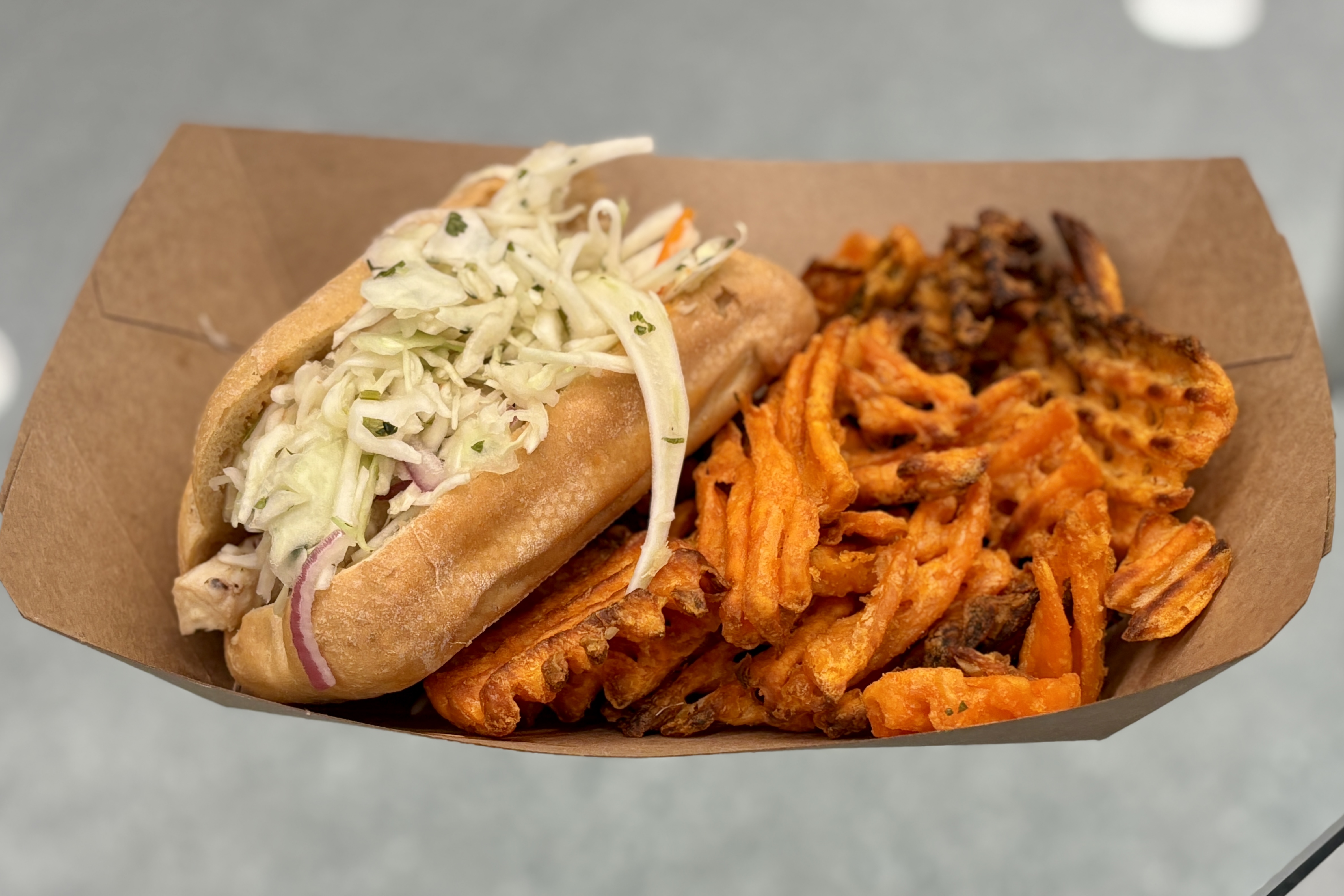 A sandwich with coleslaw and sweet potato fries are served in a cardboard container.