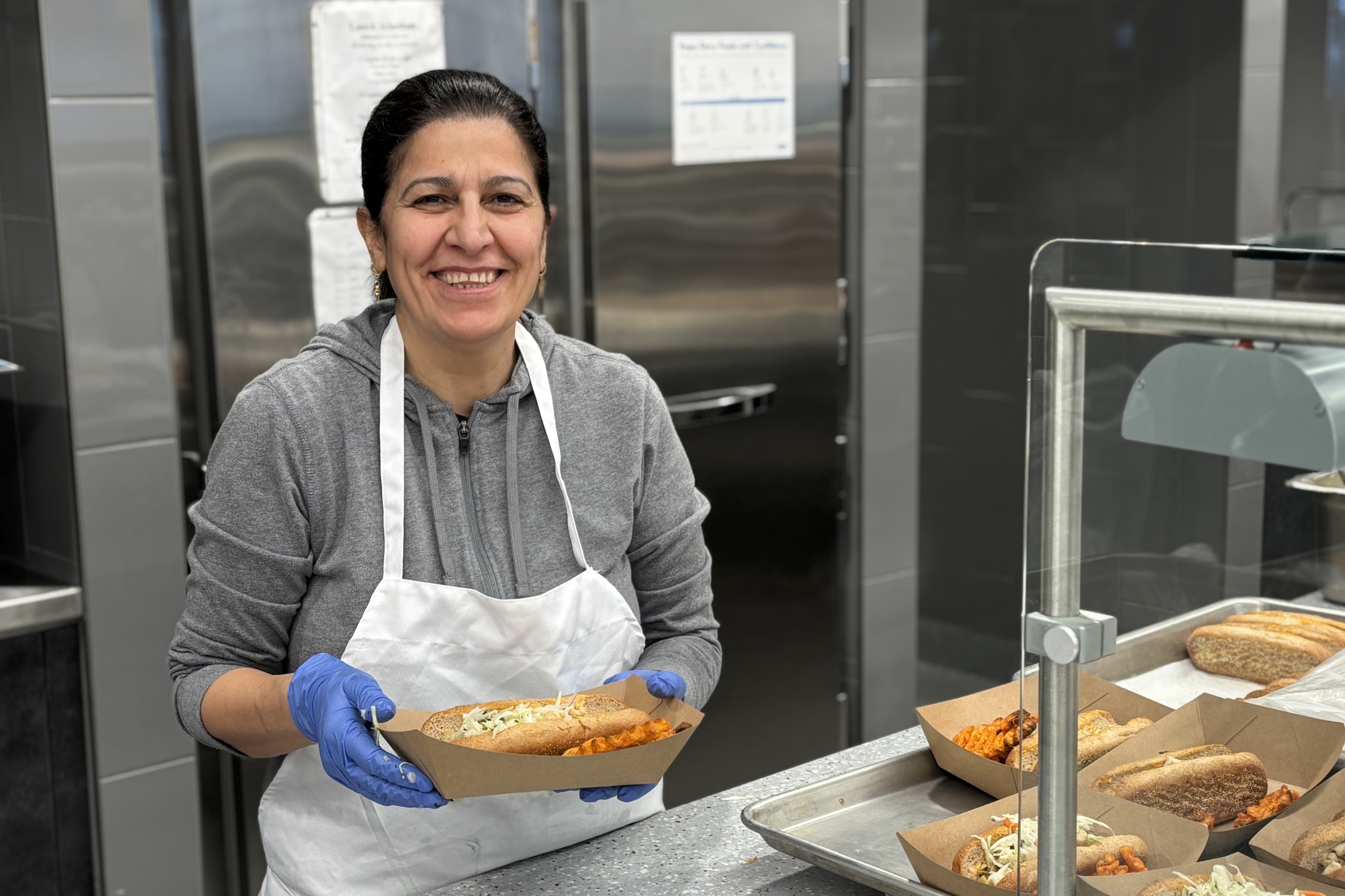 A smiling woman in an apron holds a tray of sandwiches in a commercial kitchen.