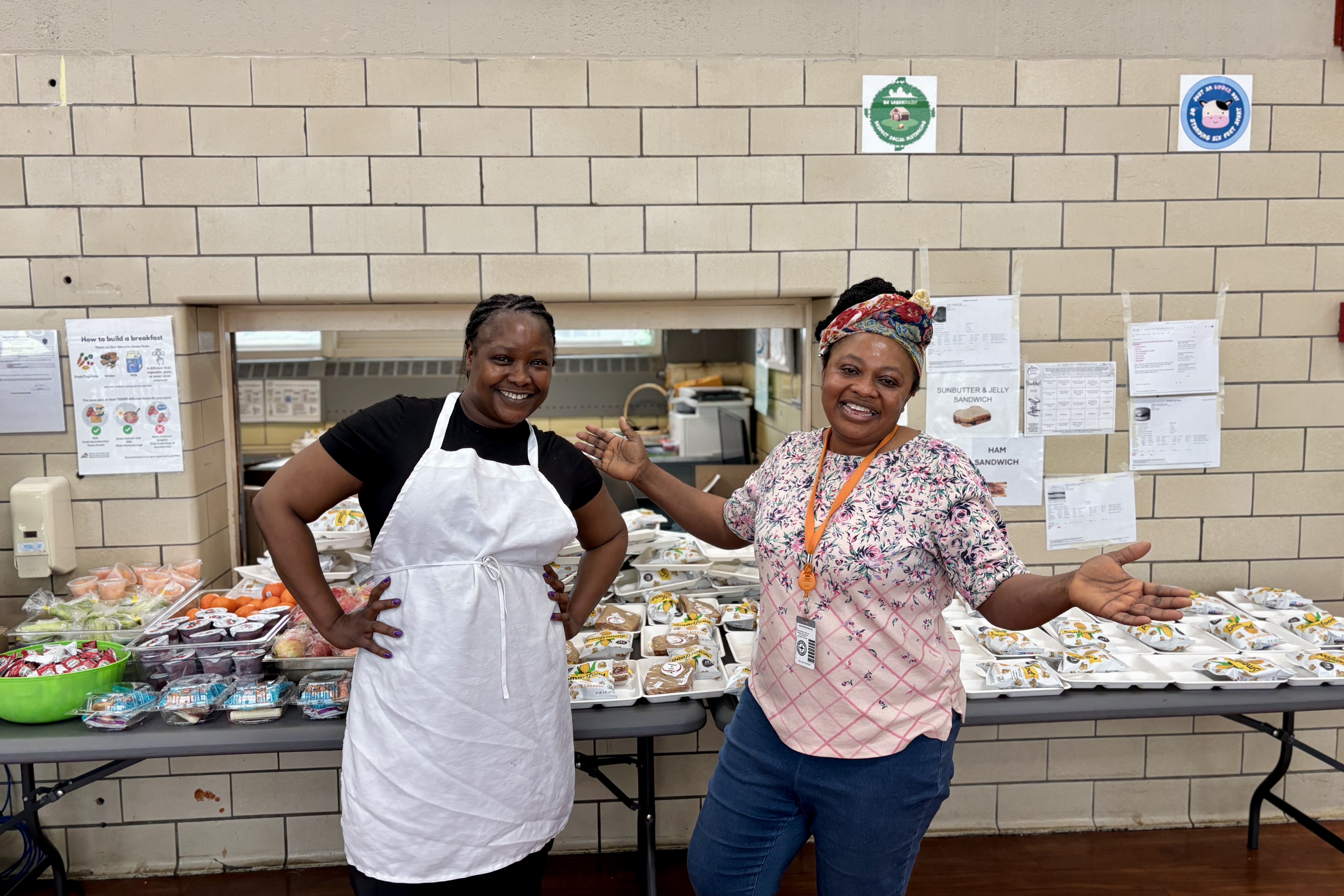 Two women smile, standing behind a table of food in a school cafeteria.