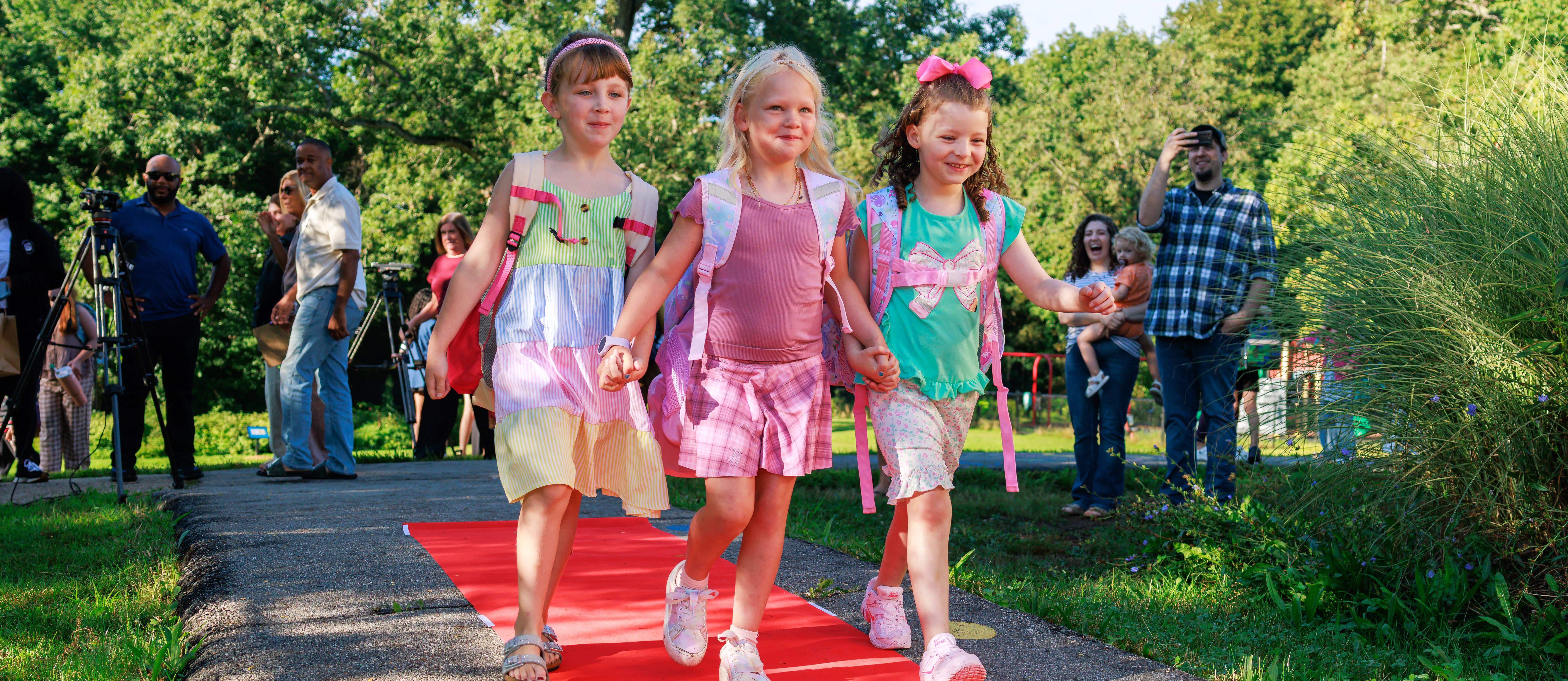 Three young students walk hand-and-hand on a red carpet during the first day of school.