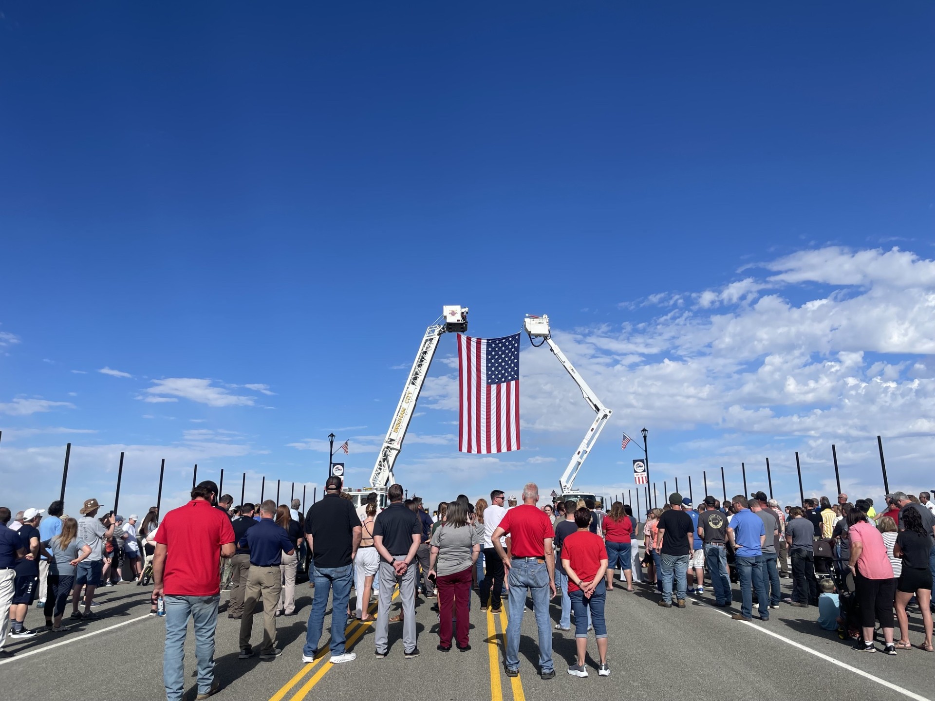 A large American flag suspended between two ladder trucks hangs above a crowd of attendees