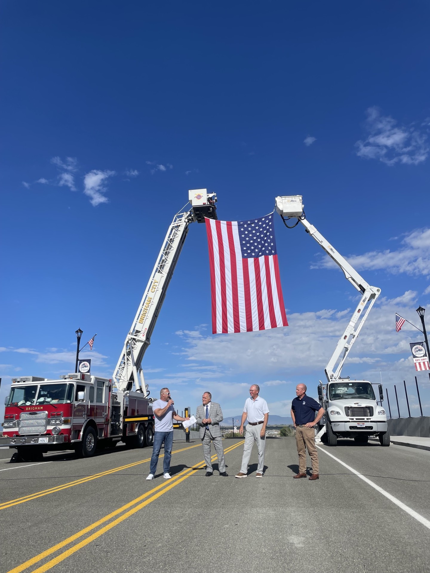 Mayor Bott, Tyler Vincent, Scott Sandall, and Joel Ferry stand below a large American flag suspended between two ladder trucks