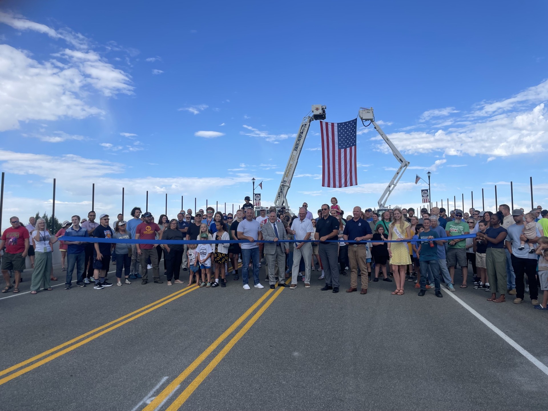 a crowd stands in front of a blue ribbon stretched across the new overpass; the man in the middle holds large scissors and is beginning to cut the ribbon