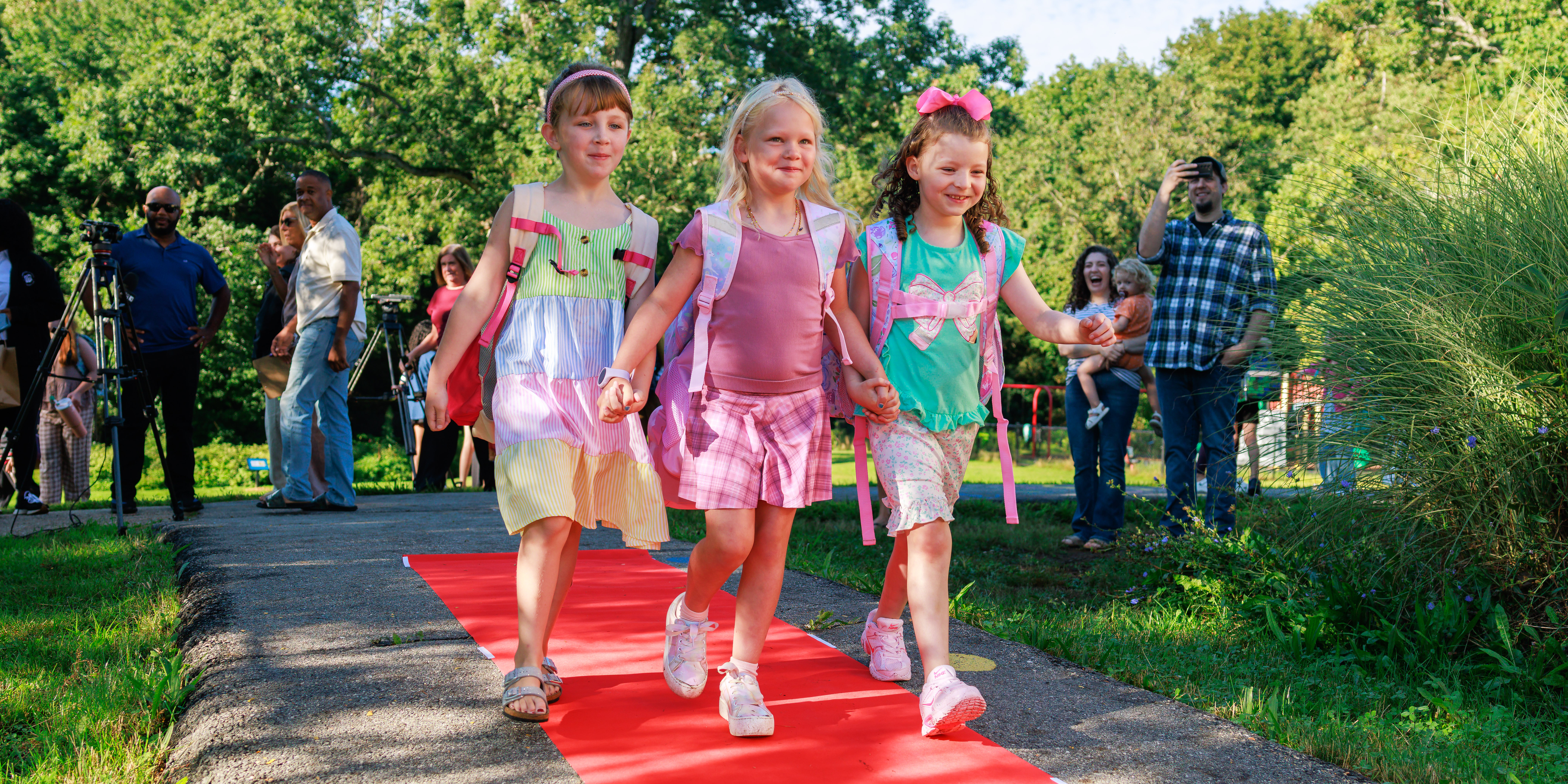 Three young students walk hand-and-hand on a red carpet during the first day of school.