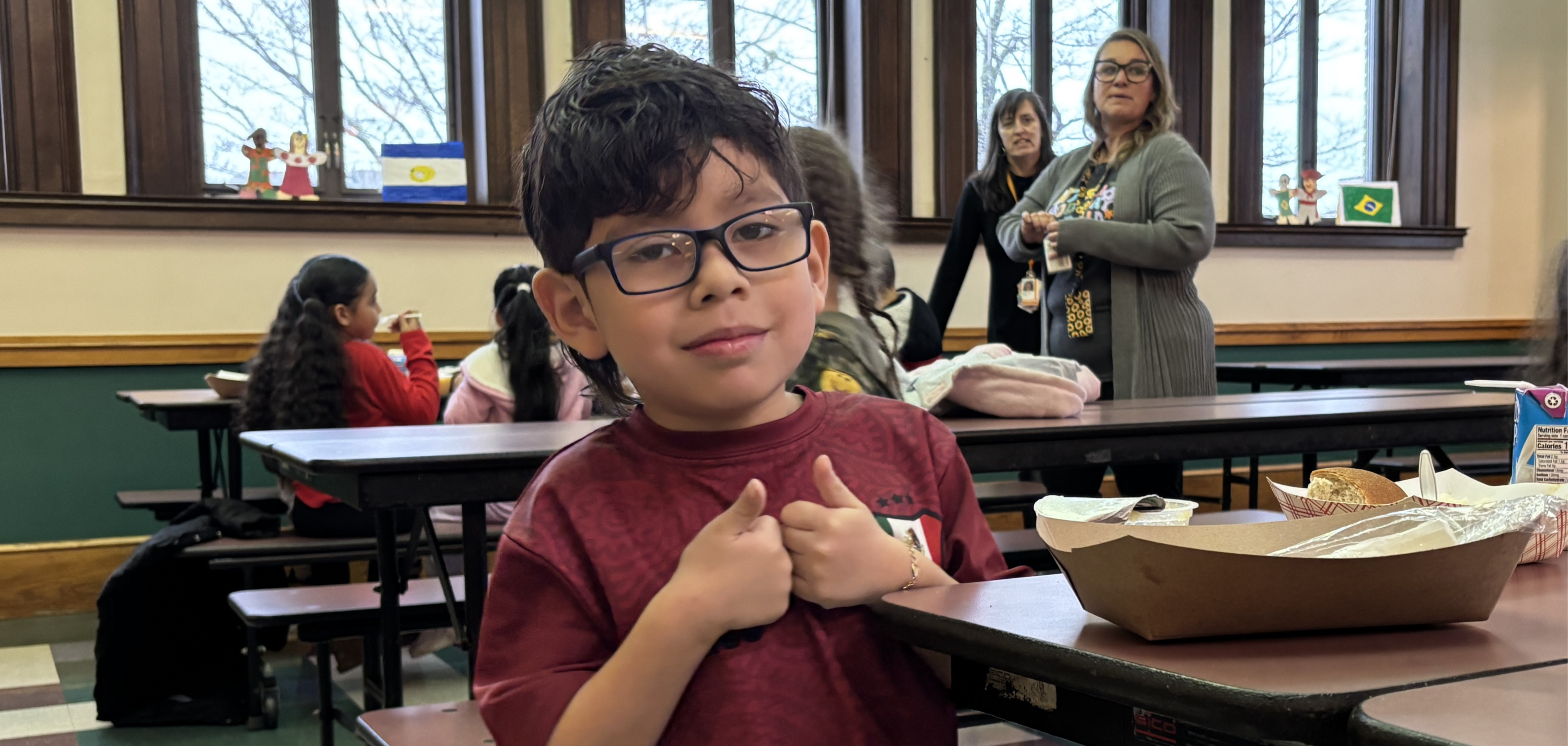 A young student wearing glasses and a maroon shirt gives two thumbs up while sitting at a cafeteria table.