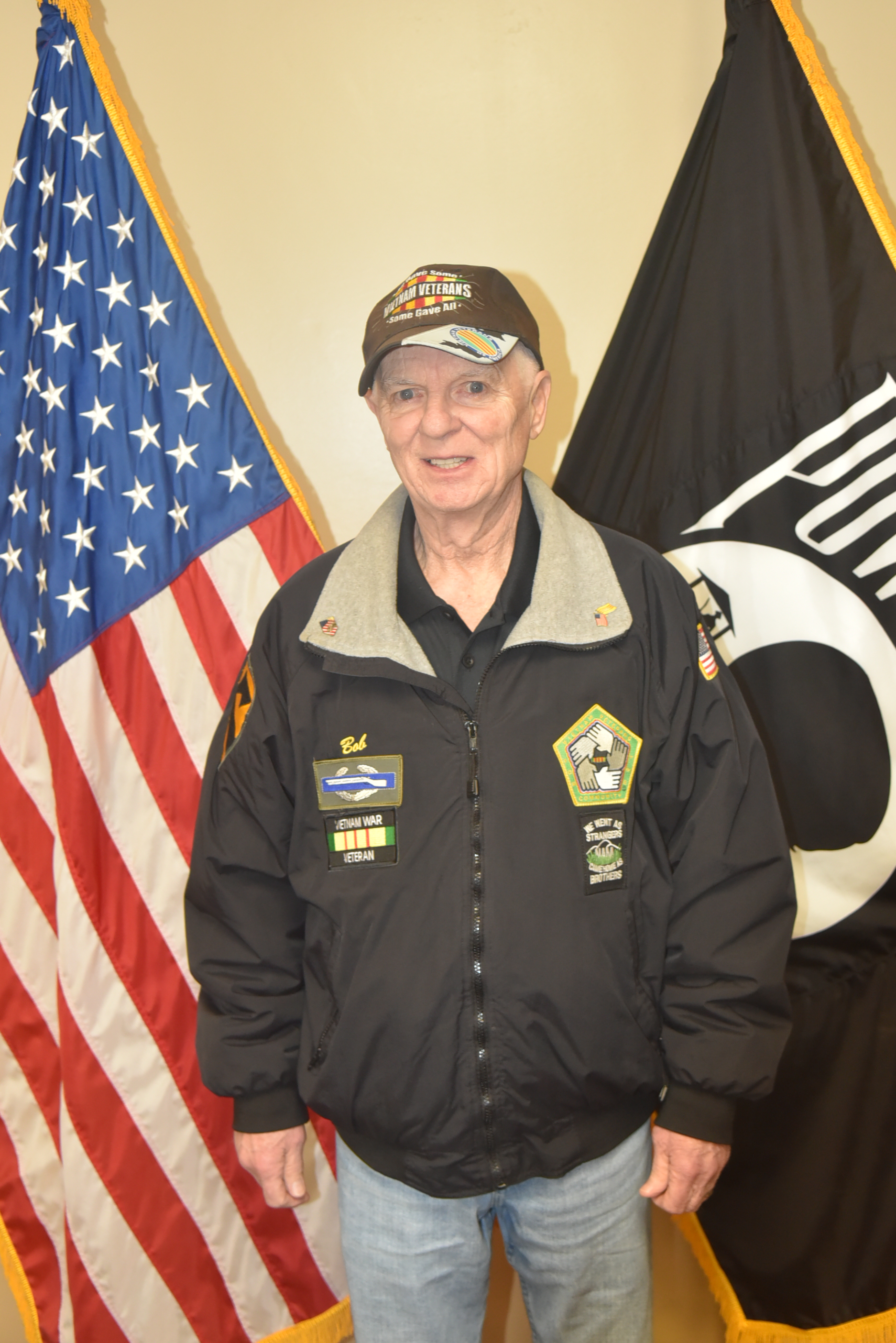 Person in black jacket and hat standing in front of two flags.