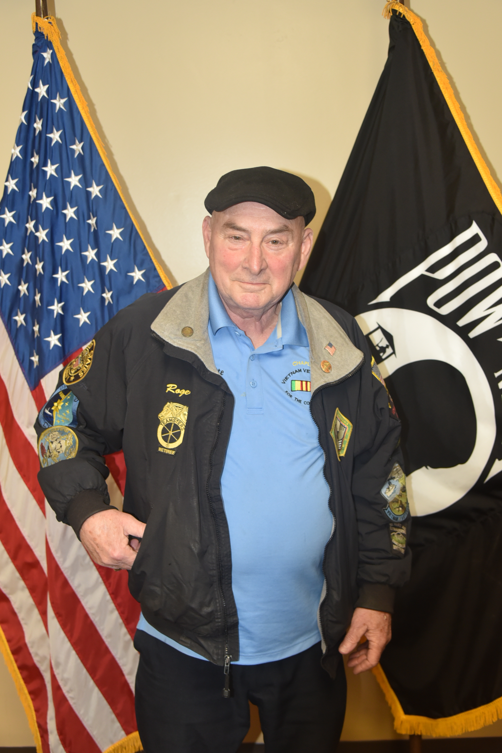 Person in blue shirt, black jacket, and black hat standing in front of two flags.