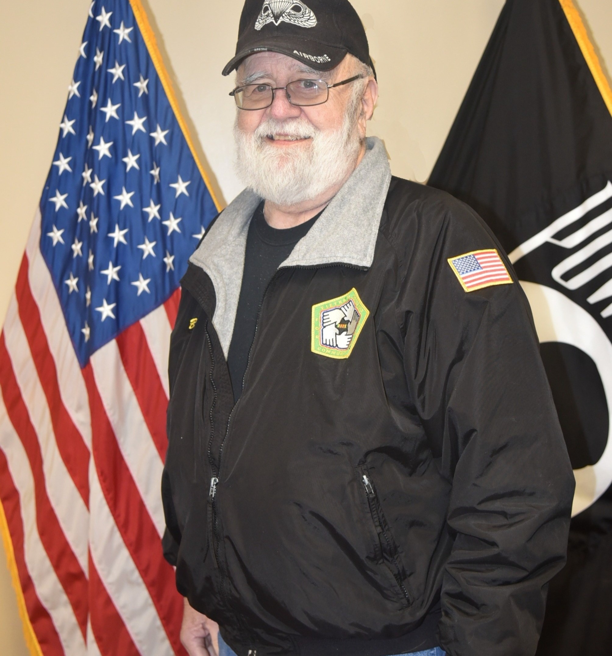 Man in black shirt, jacket, and hat standing in front of flags.