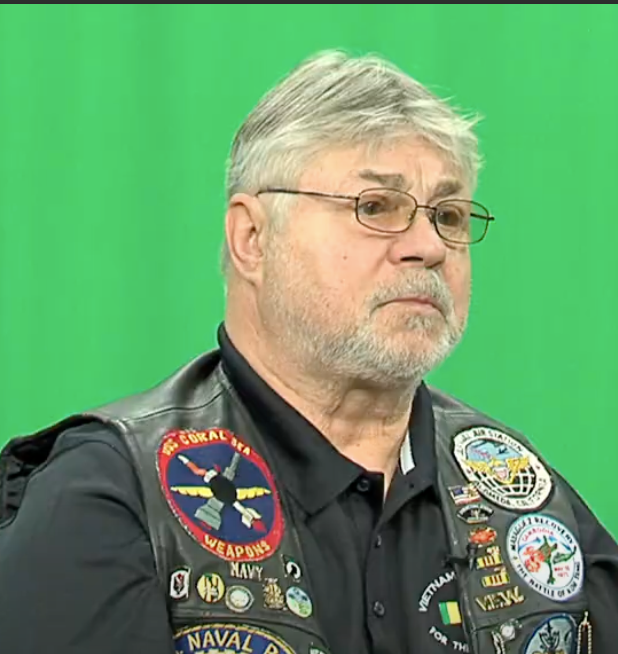 Man in front of the green screen wearing vest and glasses.