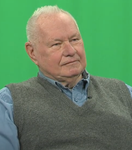 Man in front of a green screen wearing a blue button up and grey vest.