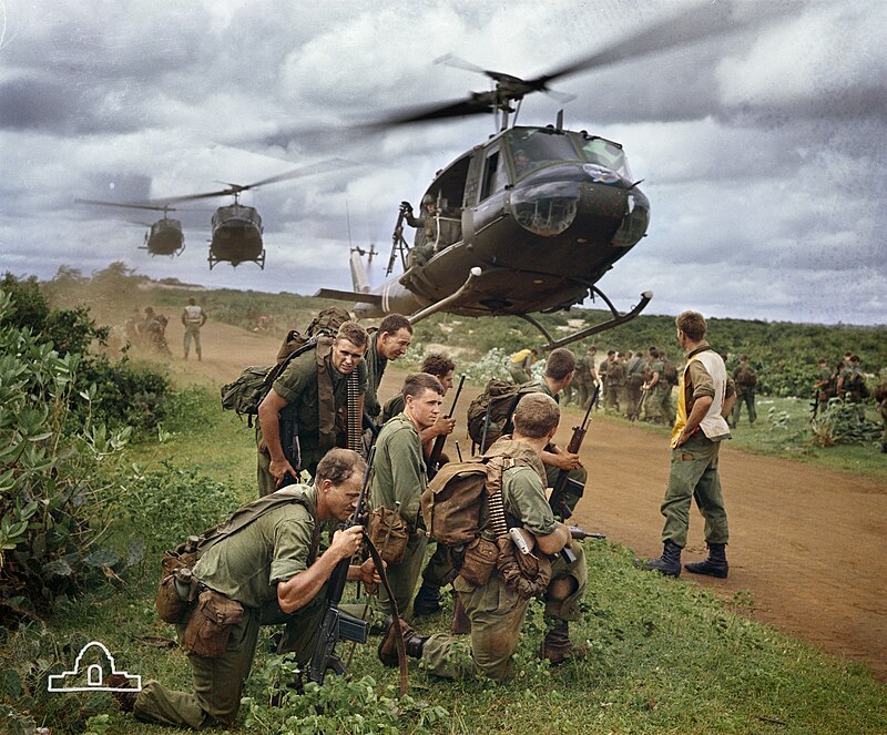 Soldiers gathered around with helicopters  landing in the middle dirt pathway.