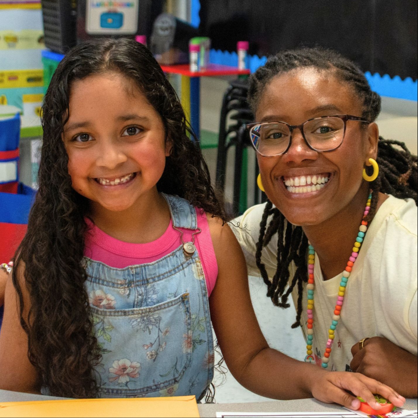 Two smiling girls at a table with a coloring sheet and a folder.