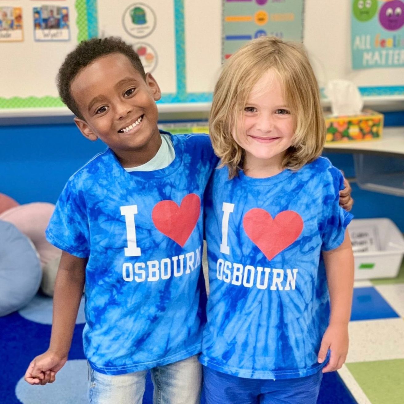 Two children in tie-dye shirts pose for a photo in a classroom, one with arms around the other.
