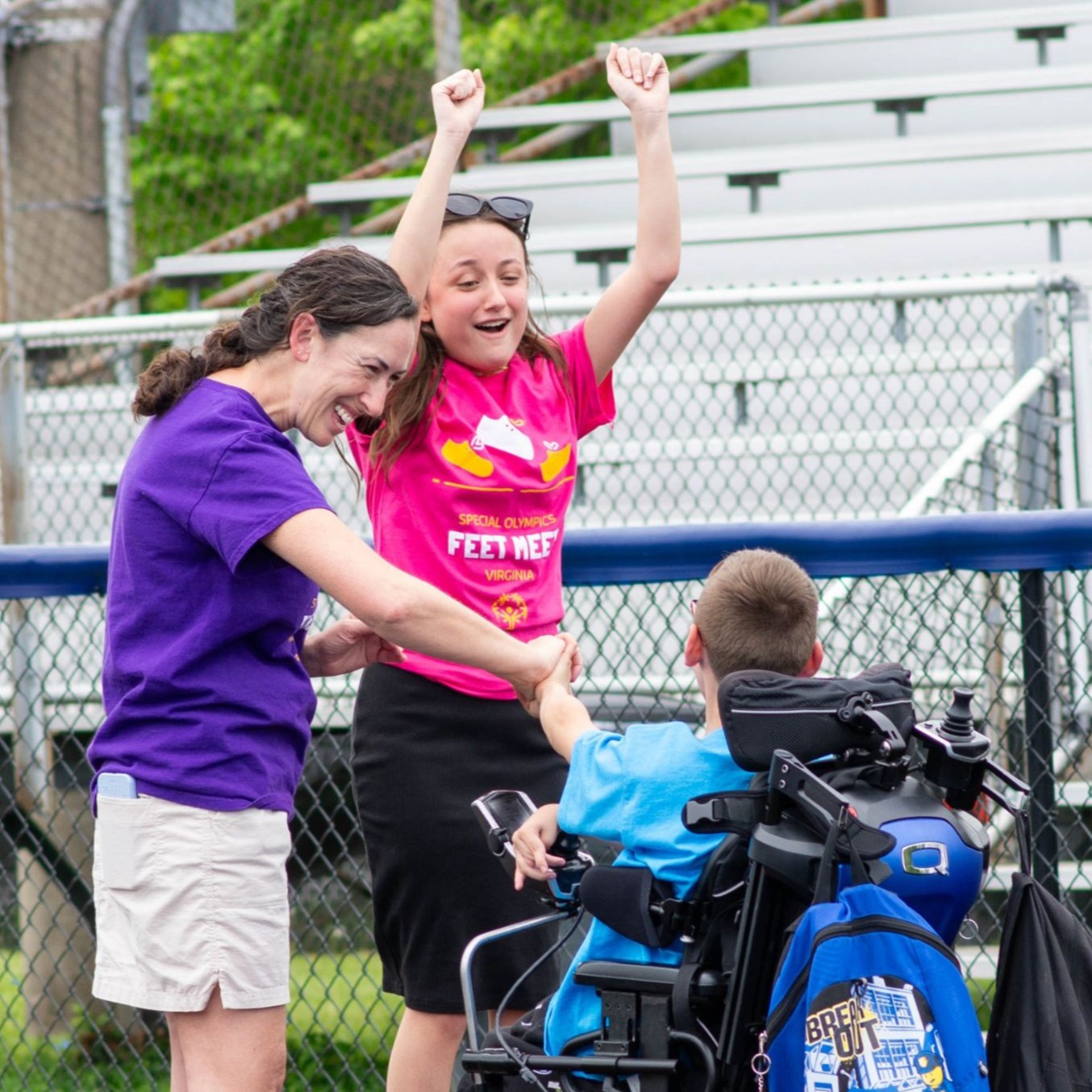 Woman and girl cheer as boy in wheelchair raises arms on a track with bleachers in the background.