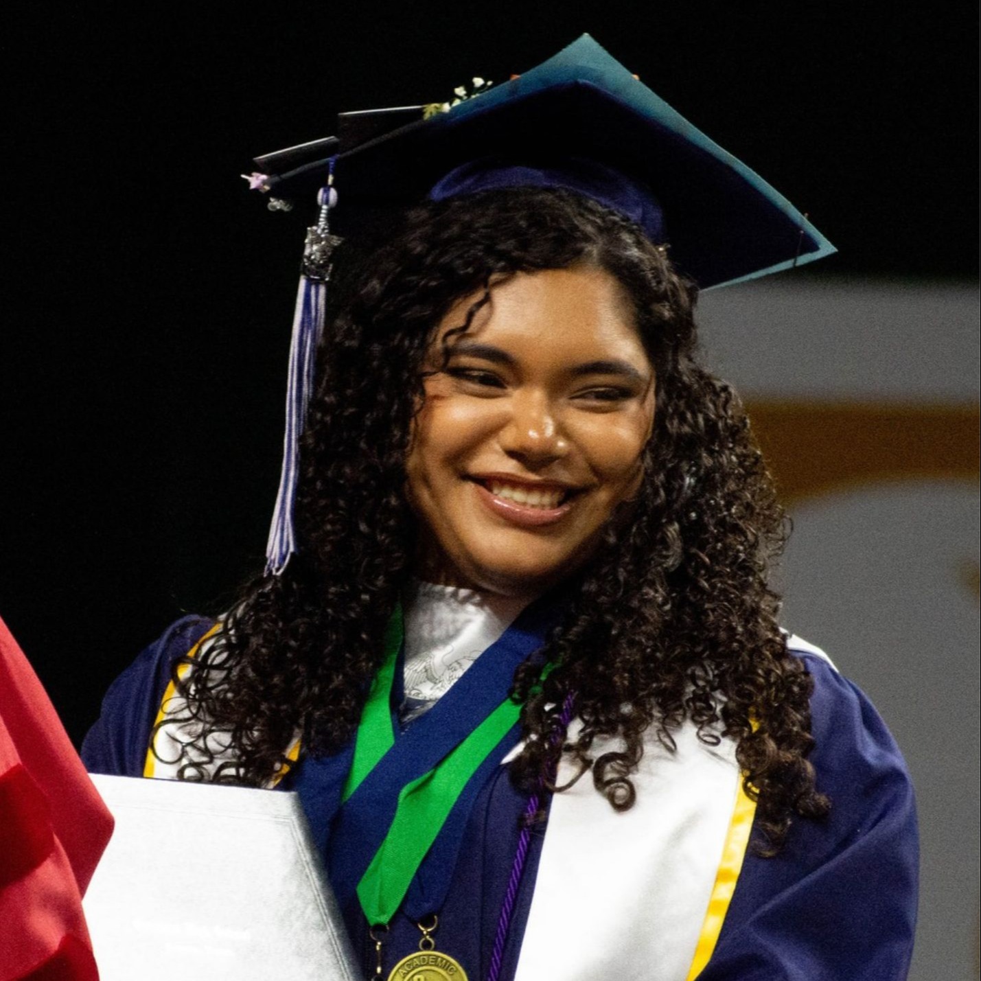 A graduate wearing cap and gown, holding a diploma, smiles while a person in red stands nearby.