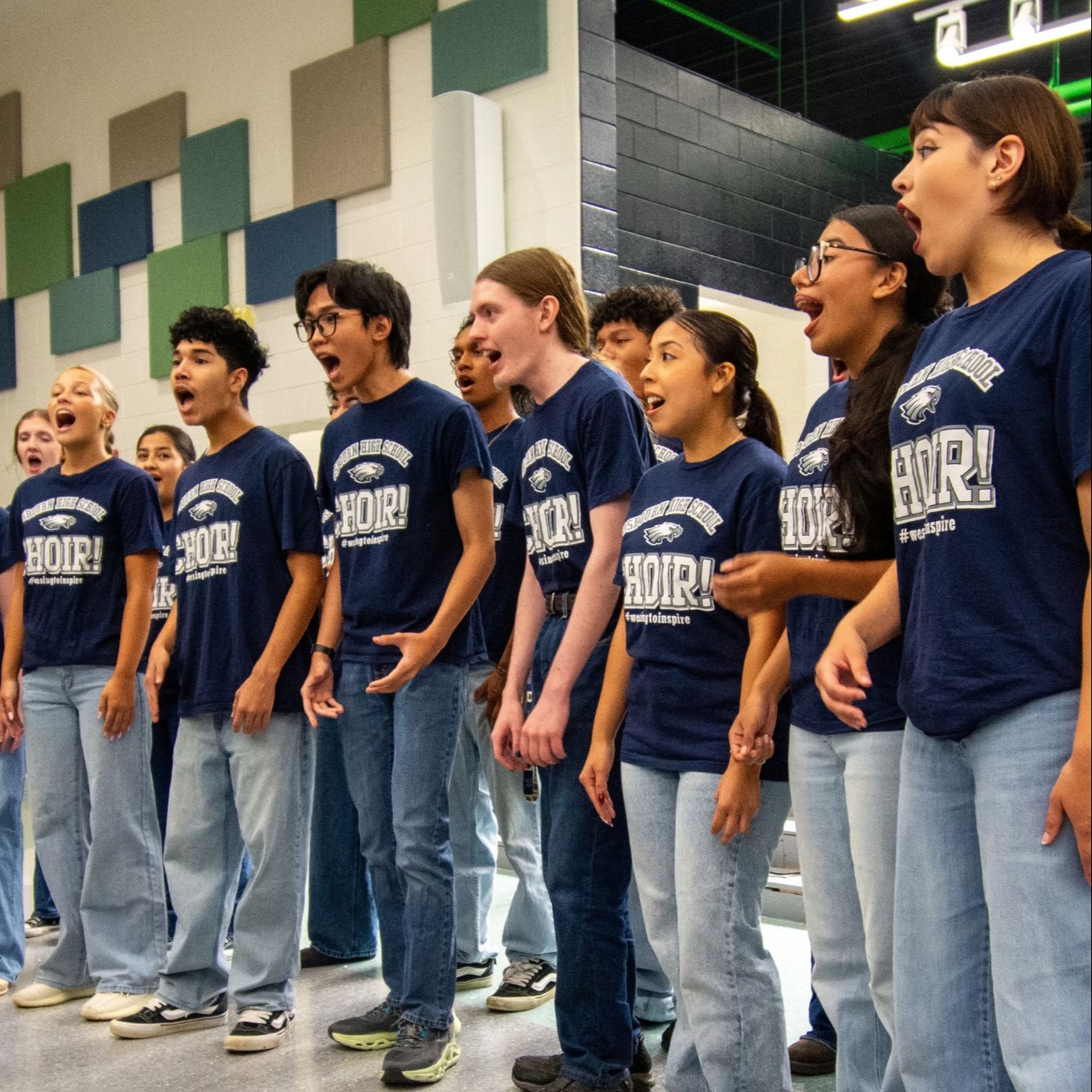Students in blue shirts and jeans stand in a row, singing in a brightly lit room with a patterned wall.