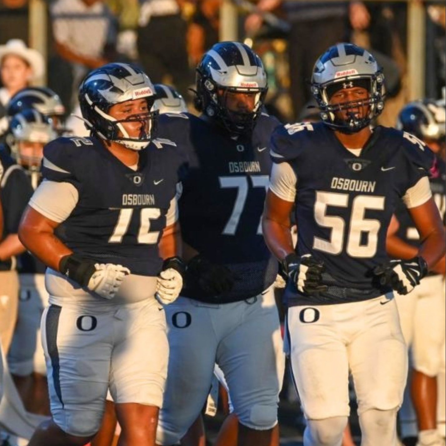 Three football players wearing navy blue jerseys, white pants, and helmets run onto a field.
