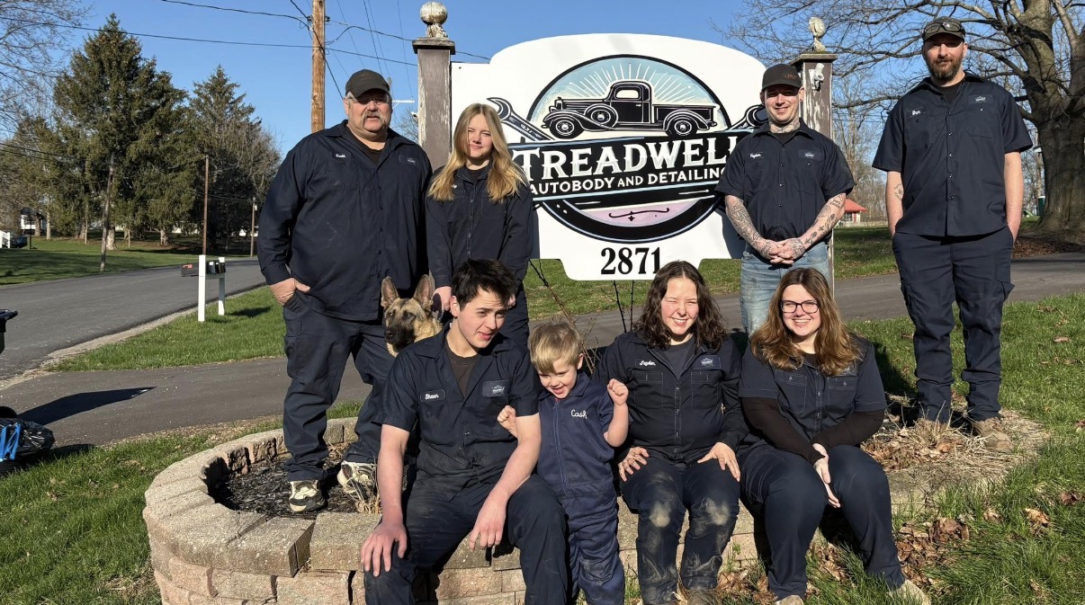 Abby and her team are posing in front of their sign in the front of their business.  
