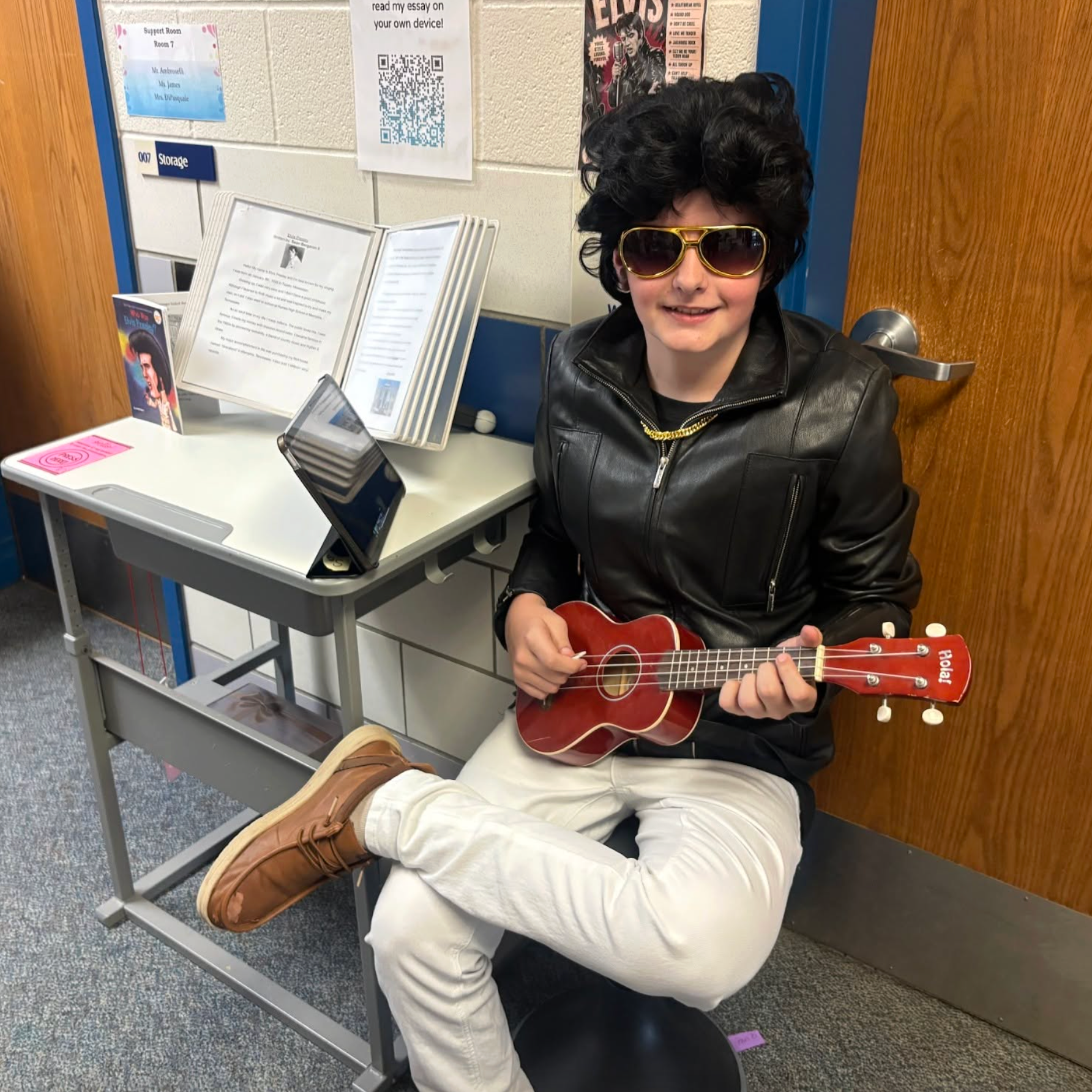 A person in Elvis-themed attire, including a wig and sunglasses, sits on a stool holding a small guitar.