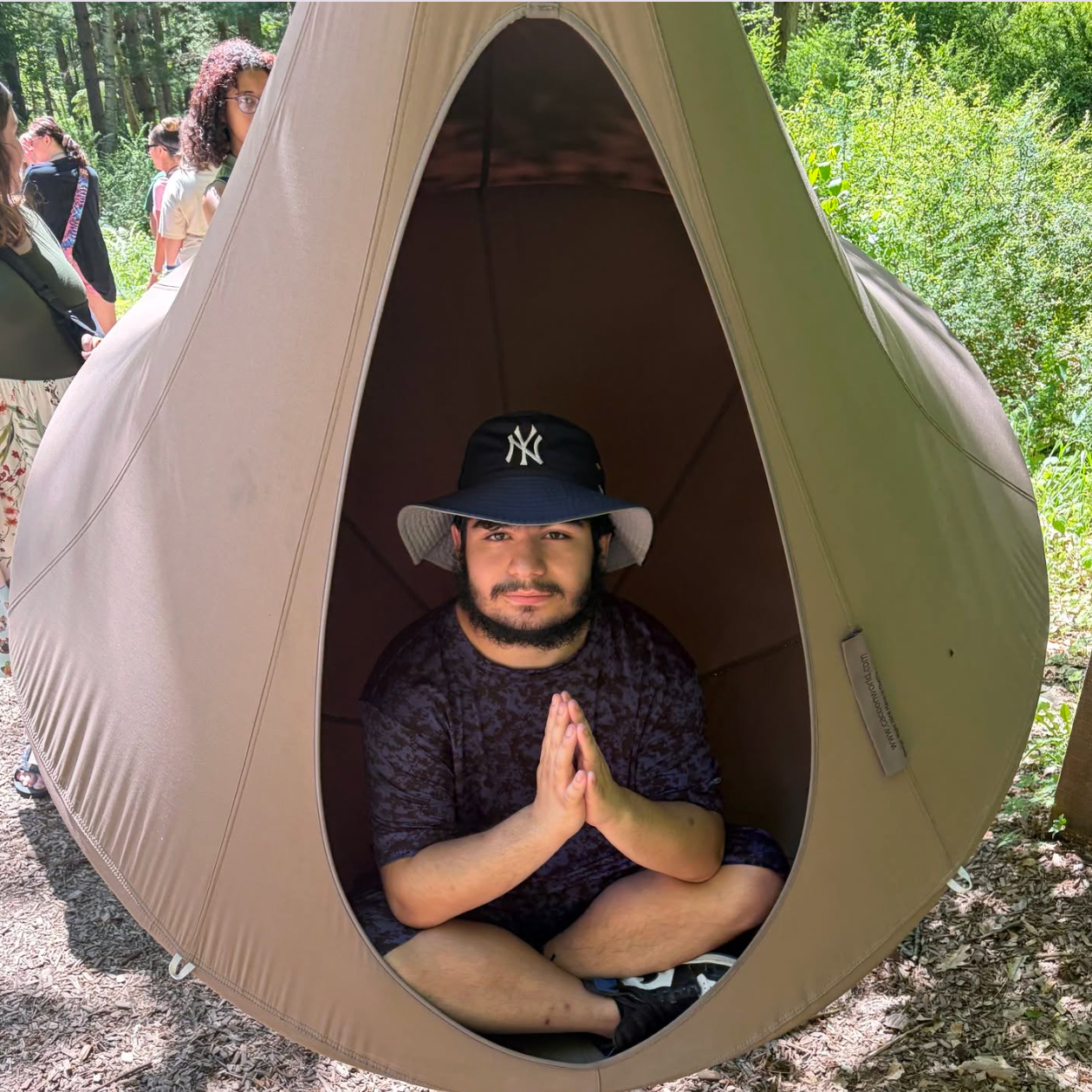 Person in a bean bag chair, sitting with hands in prayer position, with two people behind in a forest.