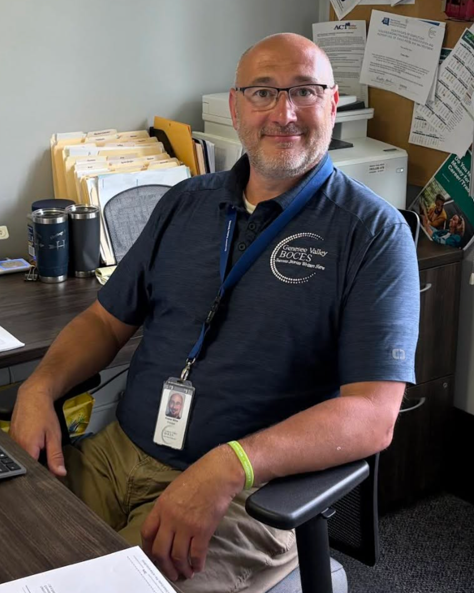 Man with glasses, ID, and lanyard sits at desk with keyboard, phone, cup, and papers.