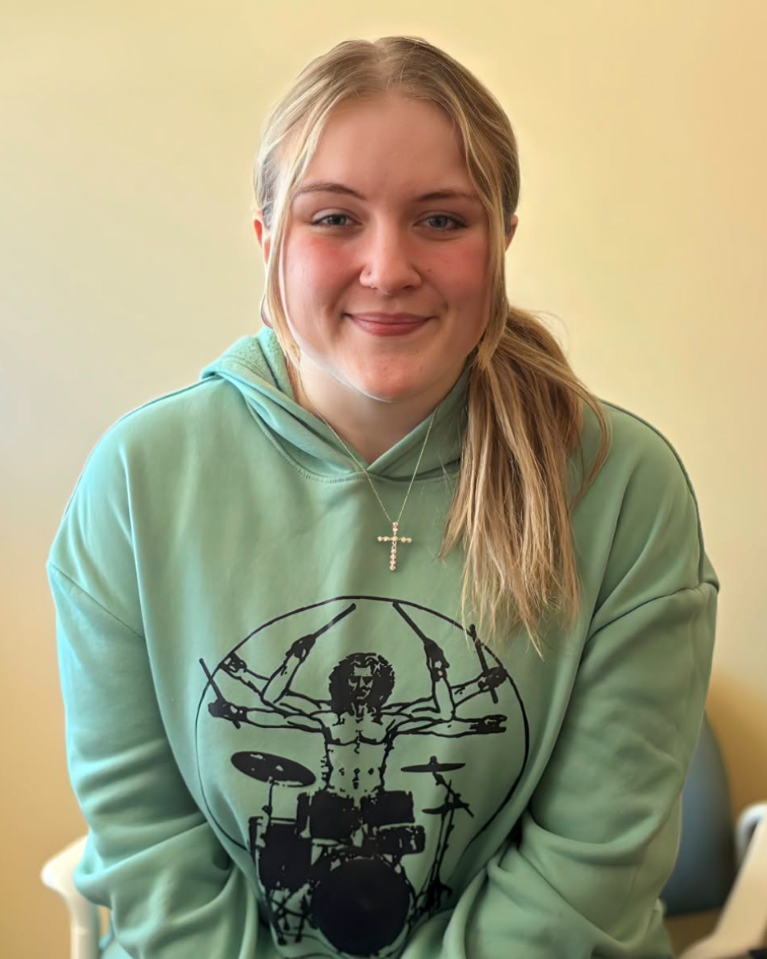 Woman with blonde hair in a green hoodie with a cross necklace sits in a chair.