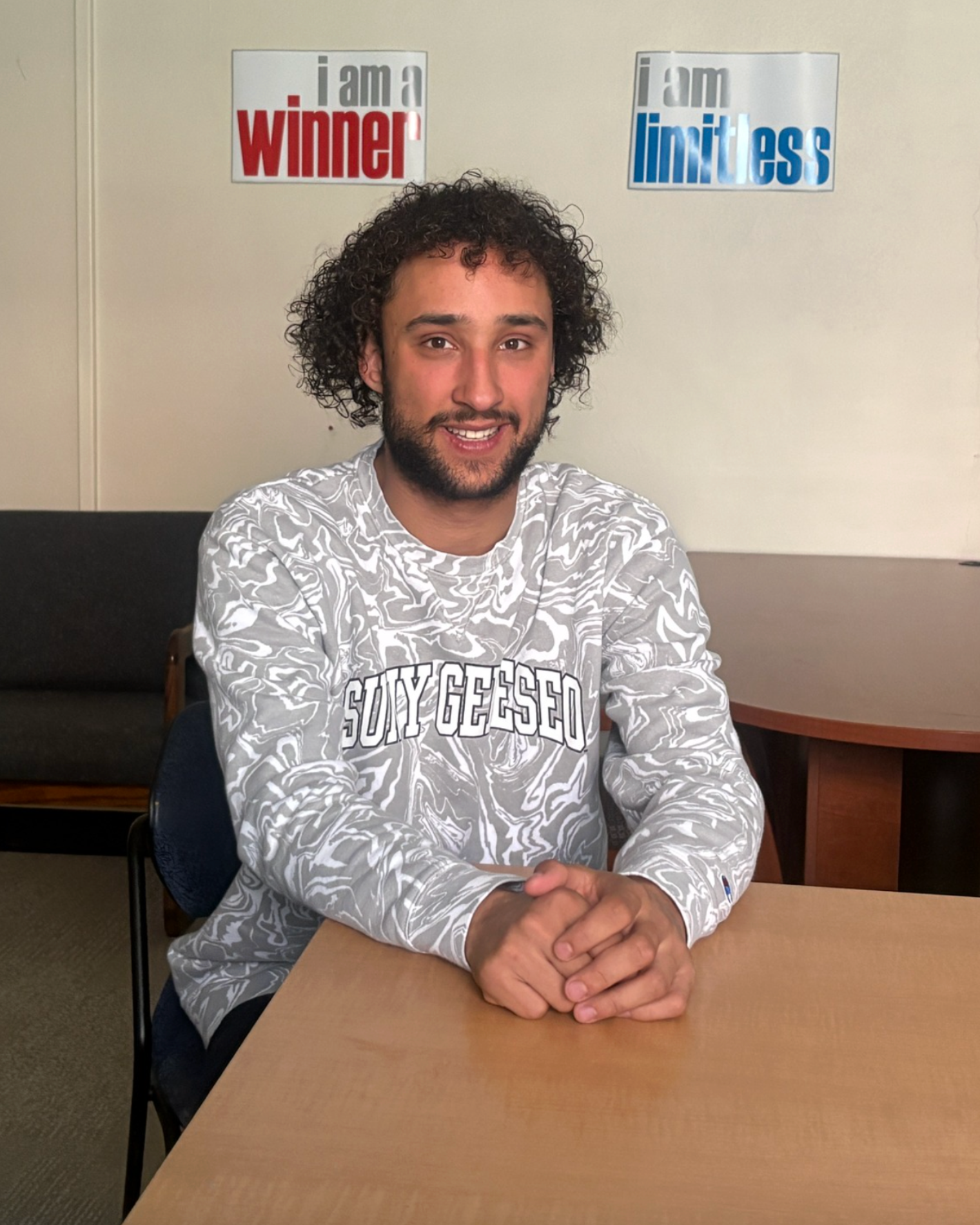 Person with curly hair in a room with chairs and motivational posters. He wears a long-sleeve shirt.